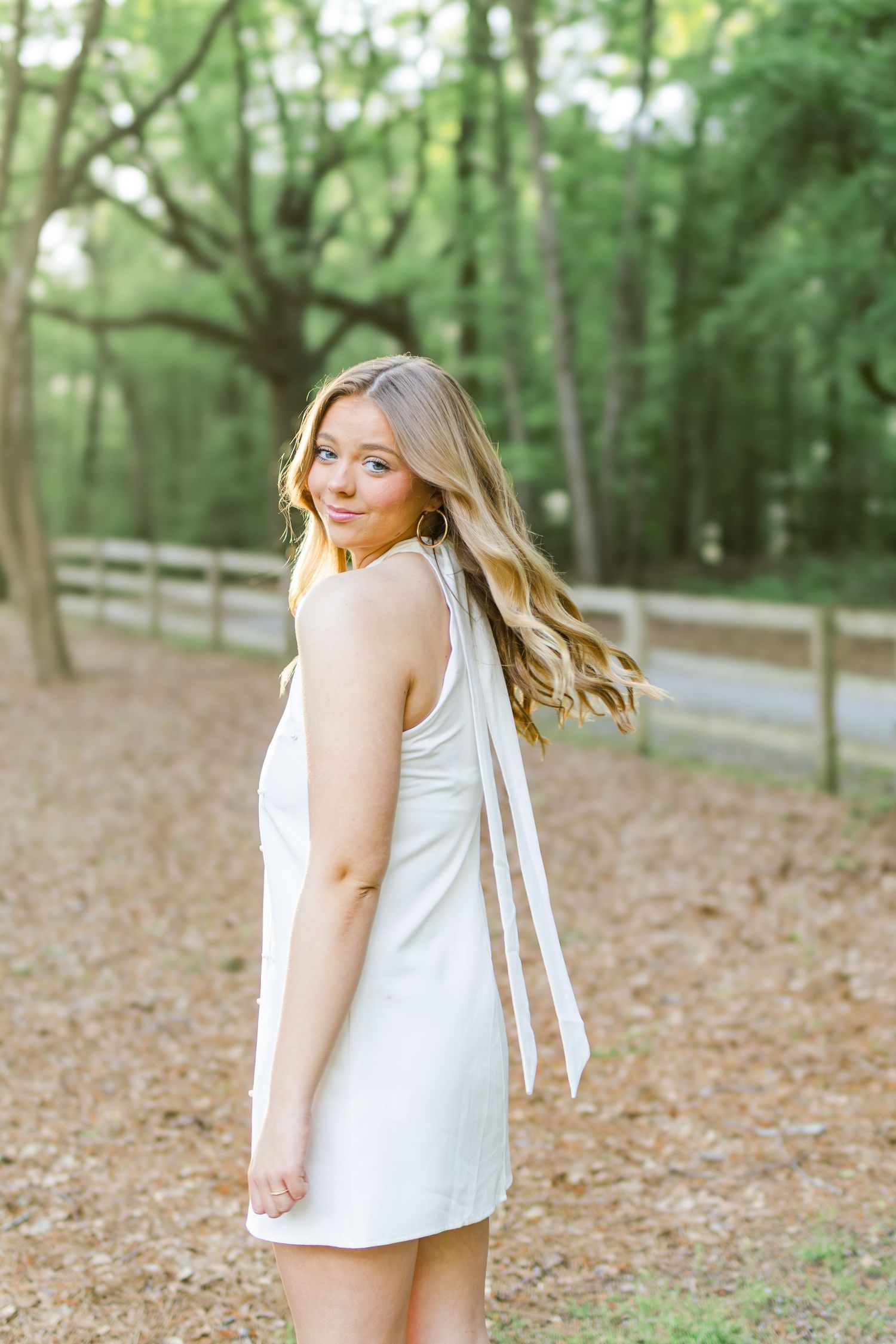 A person in a white summer dress poses on a wooded path with fallen leaves and a wooden fence in the background.