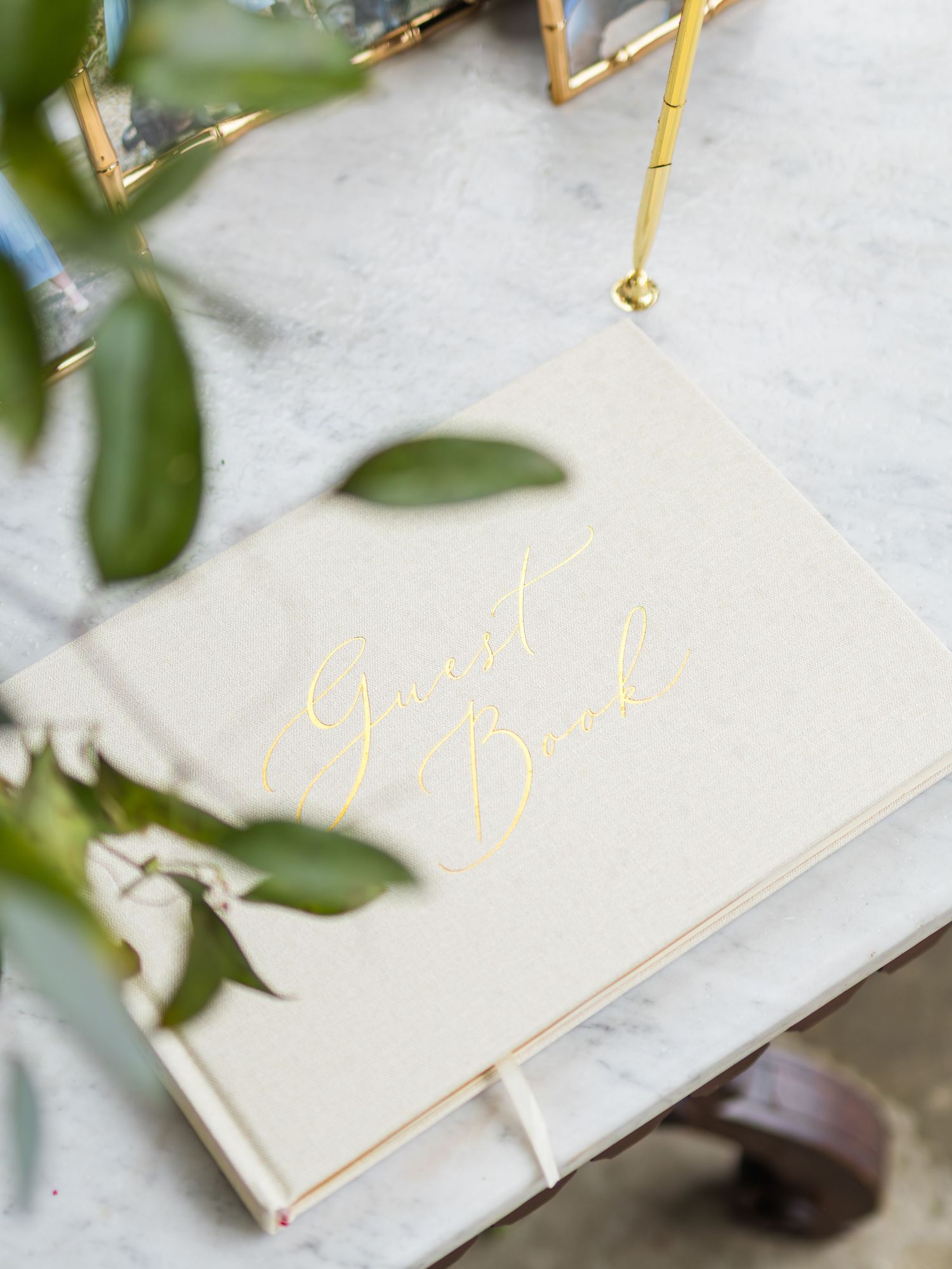 Open rustic guest book with gold lettering lies on marble surface surrounded by green leaves.