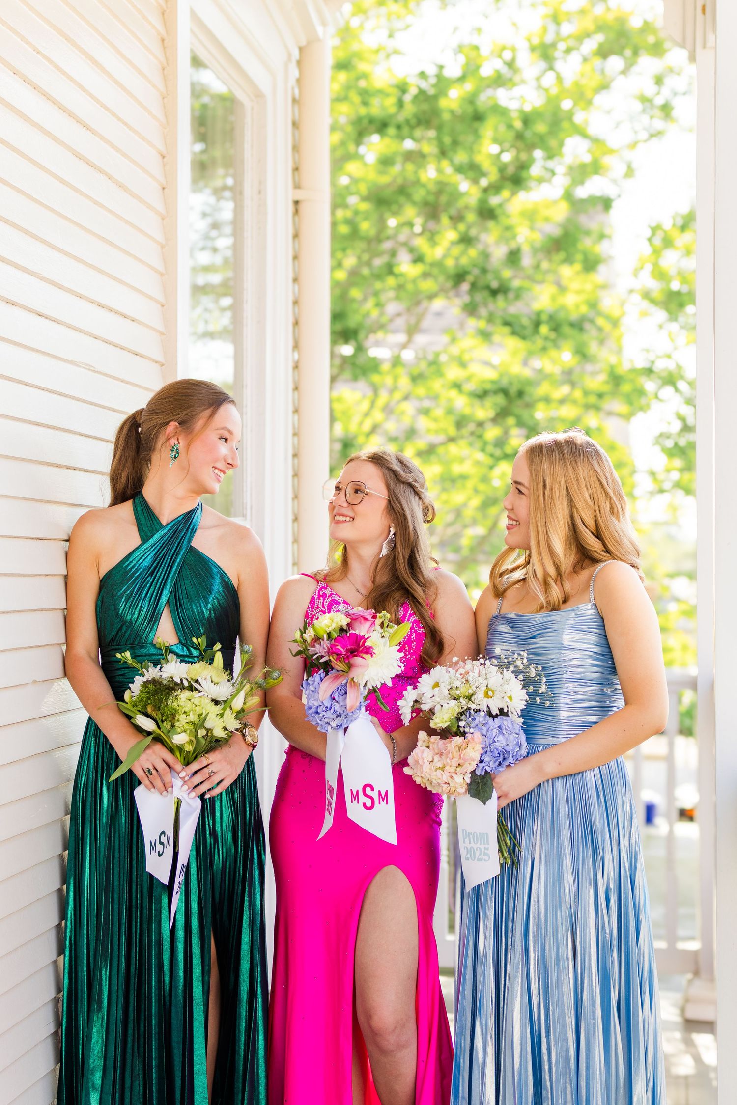 Bridesmaids wearing alternating emerald green, hot pink and light blue dresses laugh together while holding bouquets.