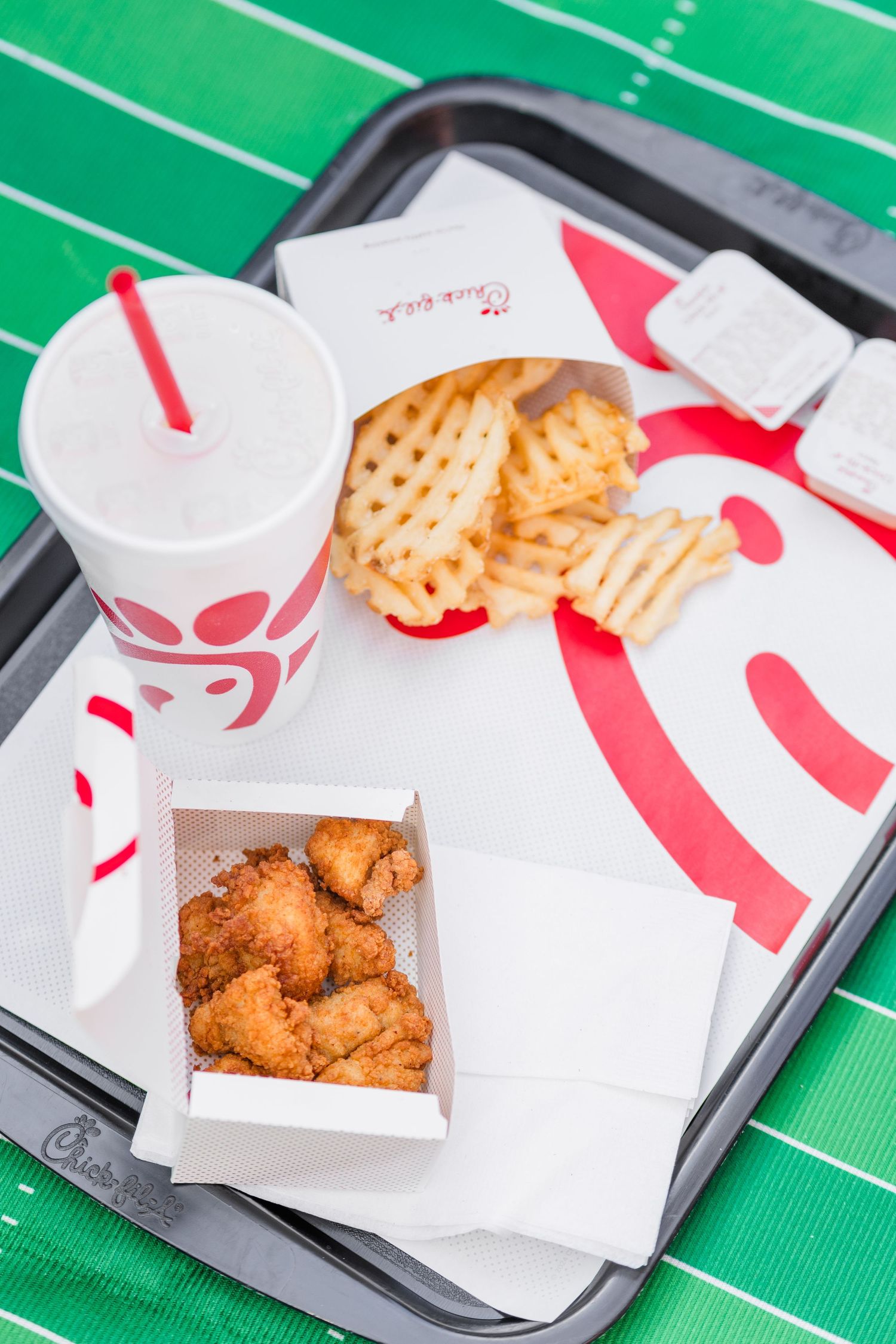 Chick-fil-A chicken nuggets and waffle fries arranged on white paper wrappers on a green football field background.