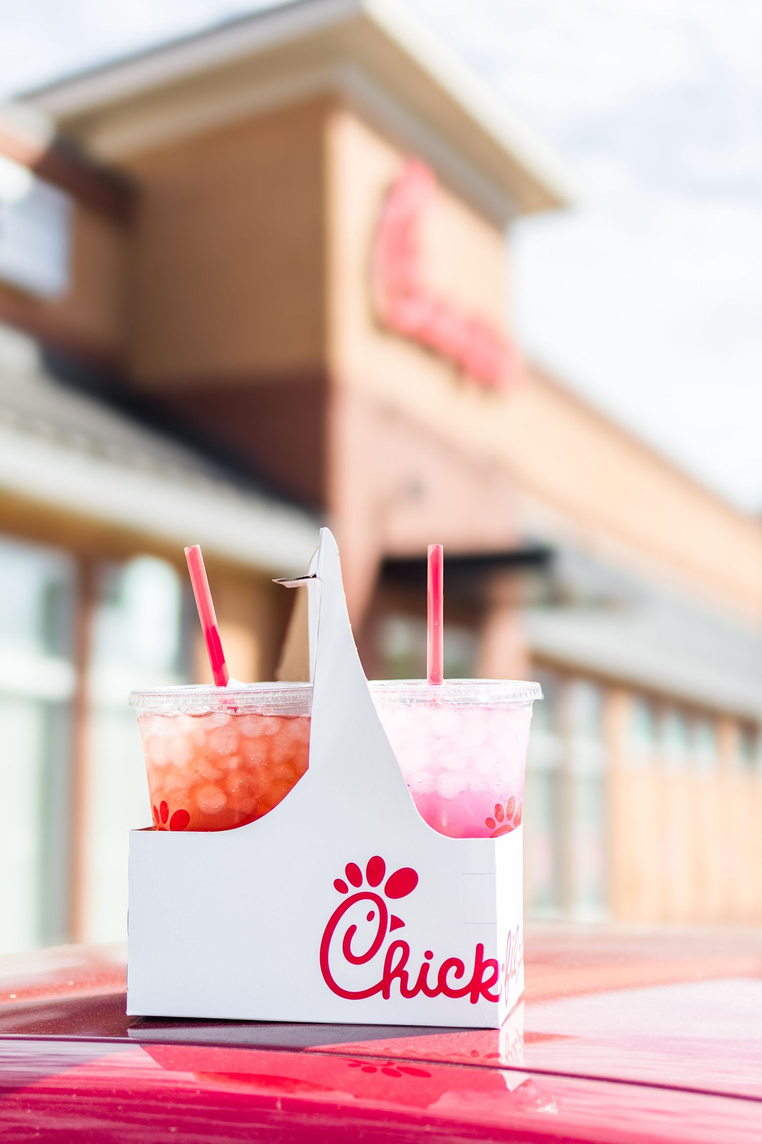 White Chick-fil-A drink carrier with pink beverages and red straws on a red surface in sunlight.