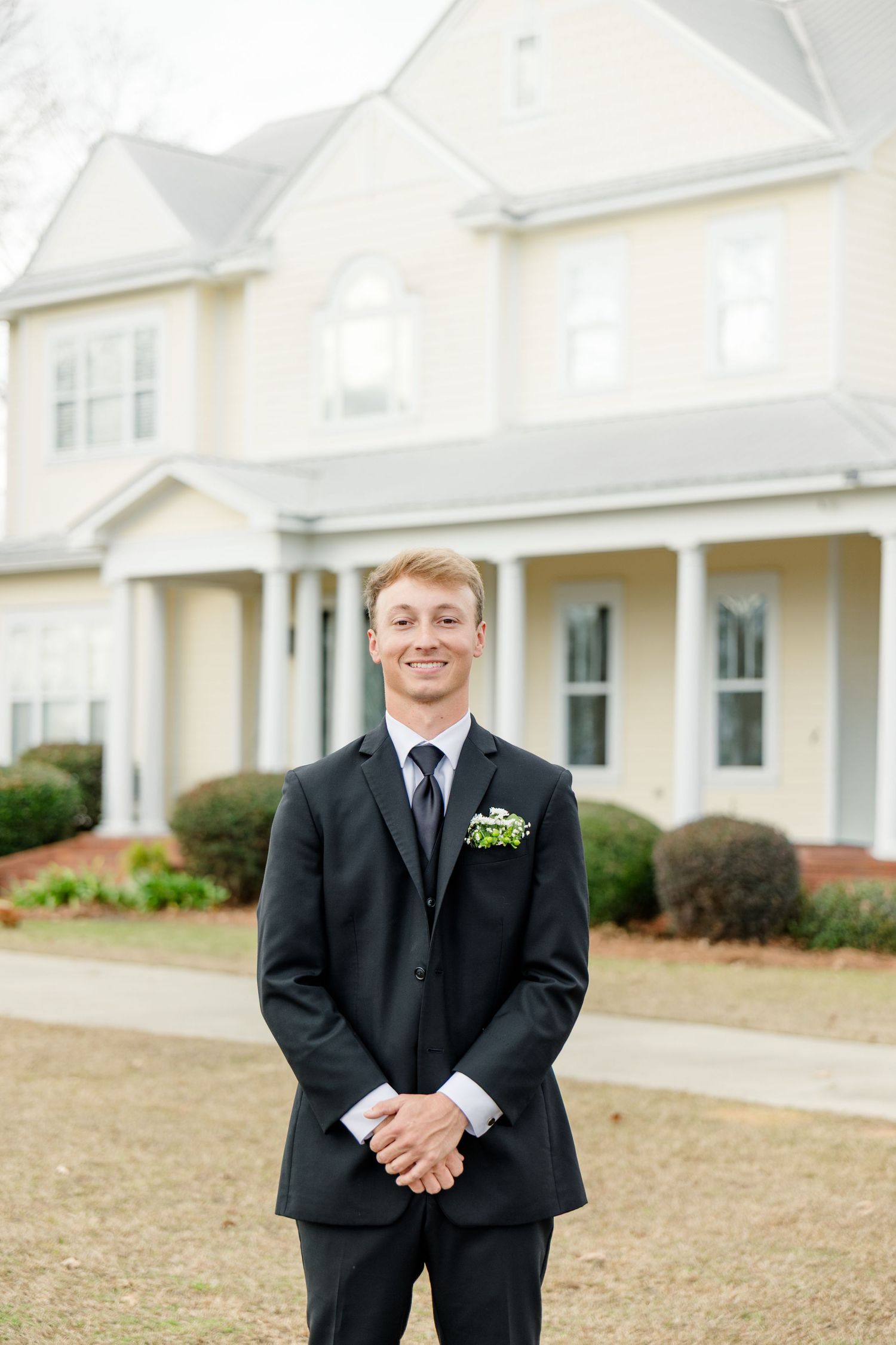 A person in a black suit stands on a lawn in front of white buildings during a formal event.