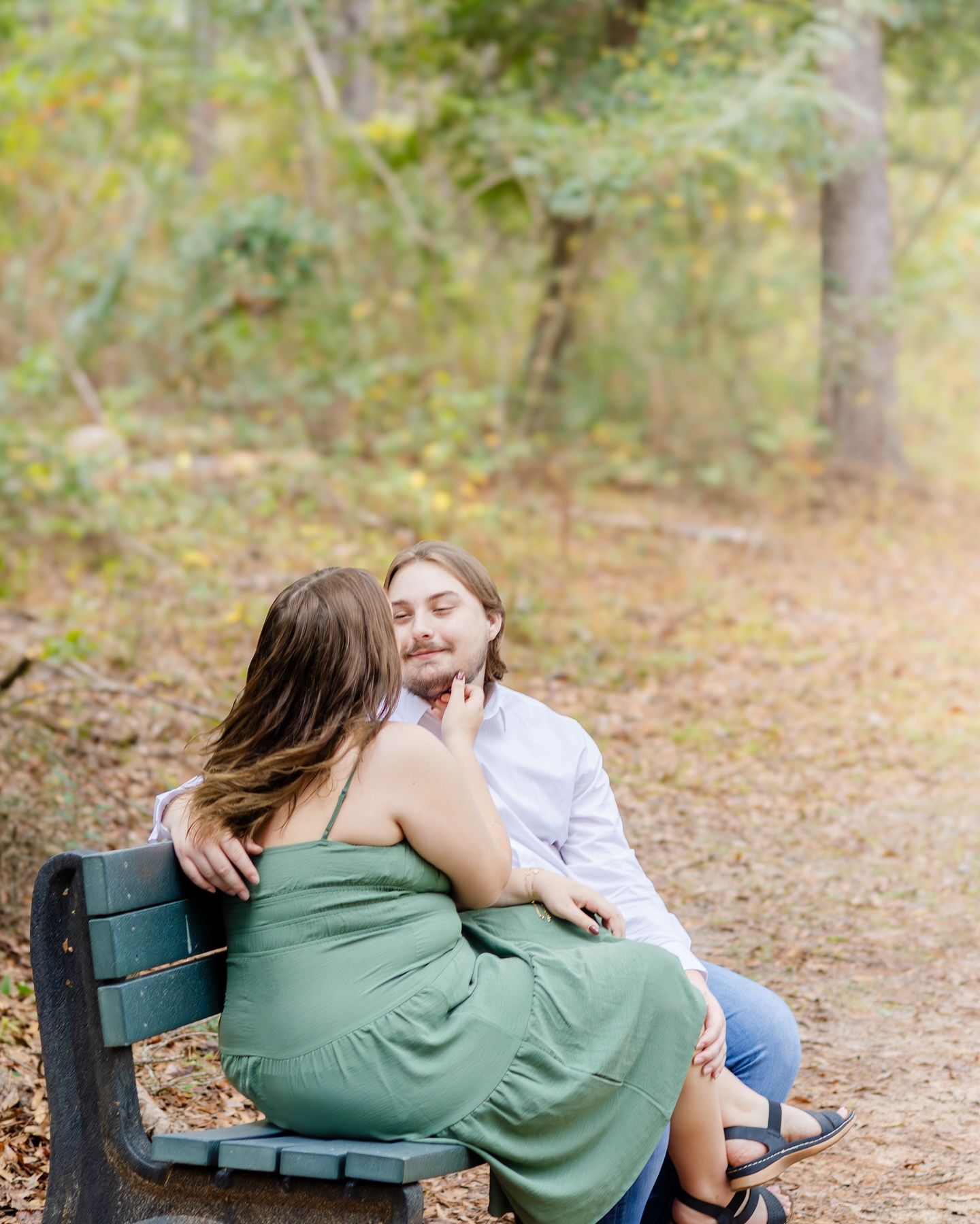 Two people share a romantic moment while sitting together on a wooden bench along a sunlit forest path.