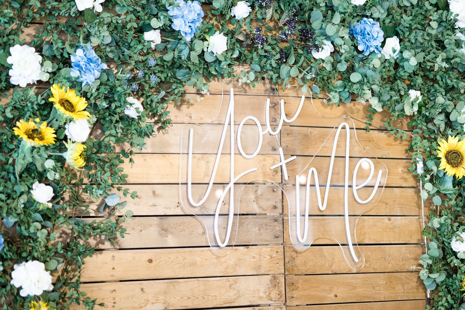 Rustic wooden wall decorated with green ivy garland, white and yellow flowers, and a neon-style love sign.