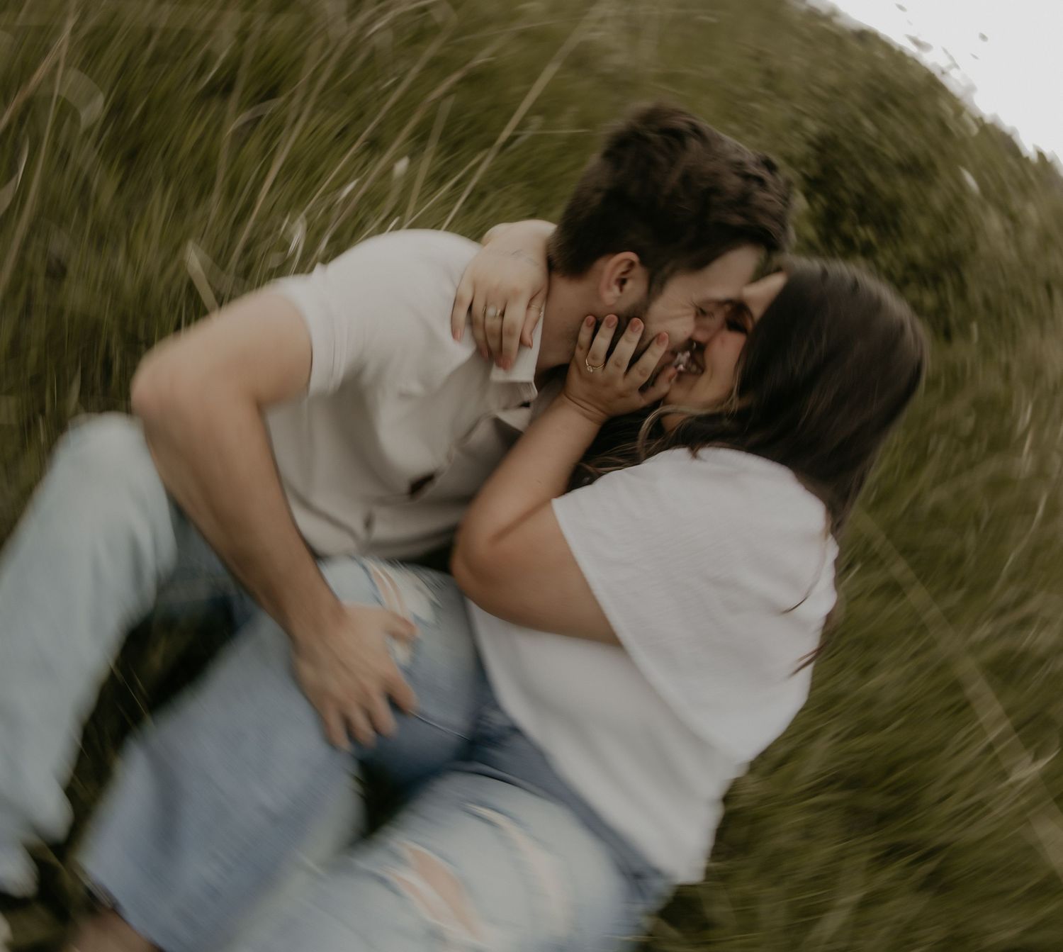 A couple lying in long grass share an intimate kiss while wearing casual clothing and embracing each other gently.
