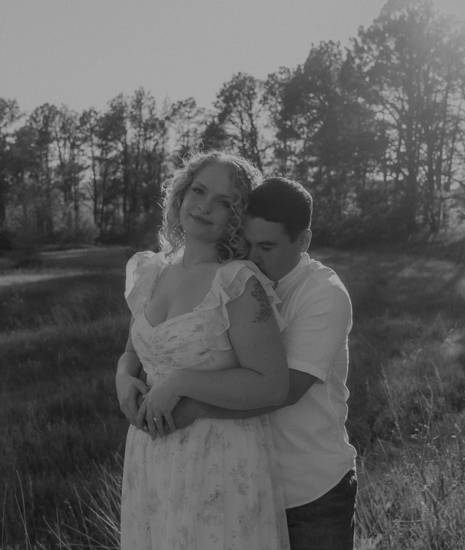A couple embraces outdoors in black and white photography with tall grass and trees in the background.