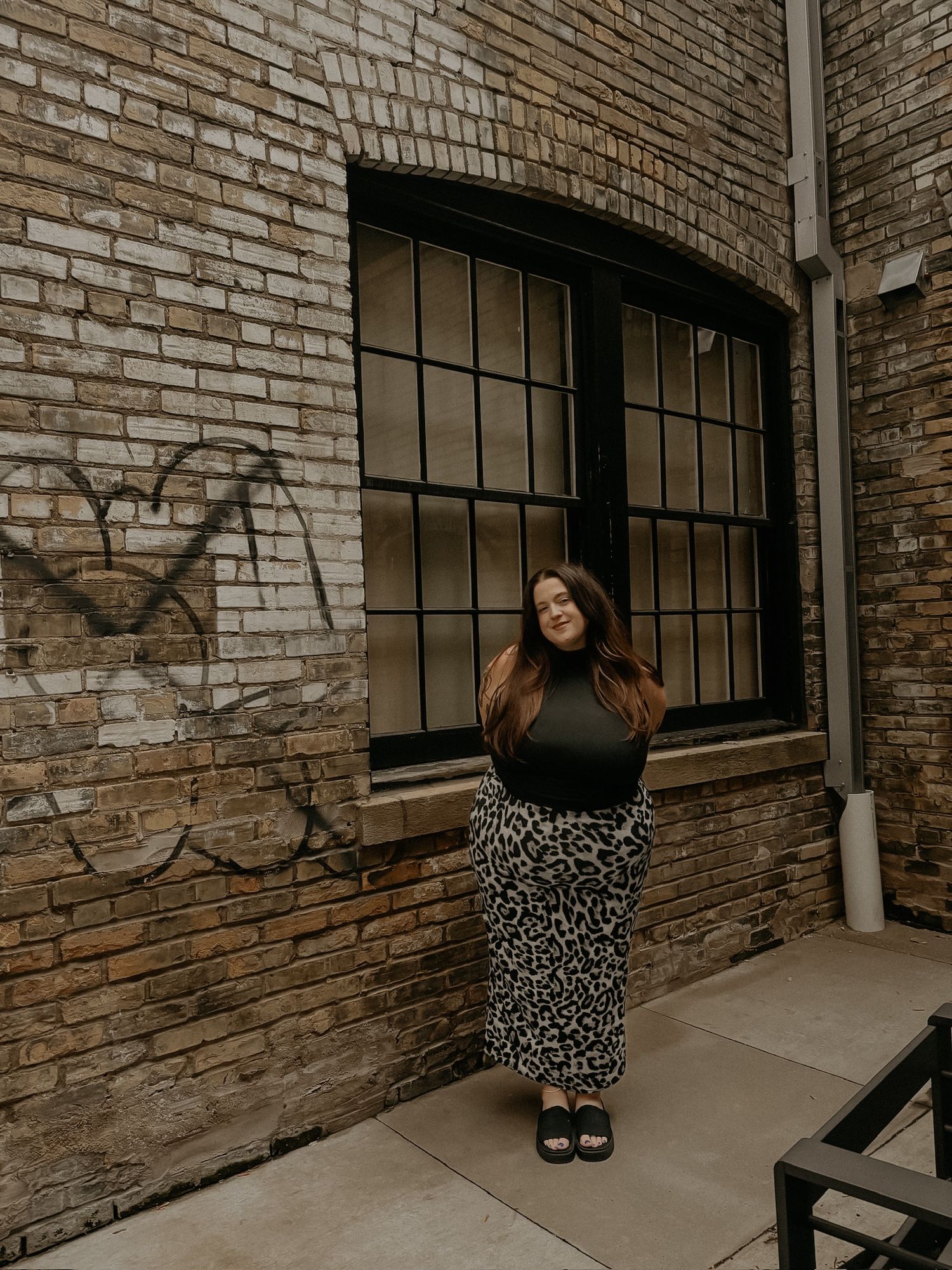 Someone wearing a black top and leopard print maxi skirt poses near a brick wall with industrial windows.