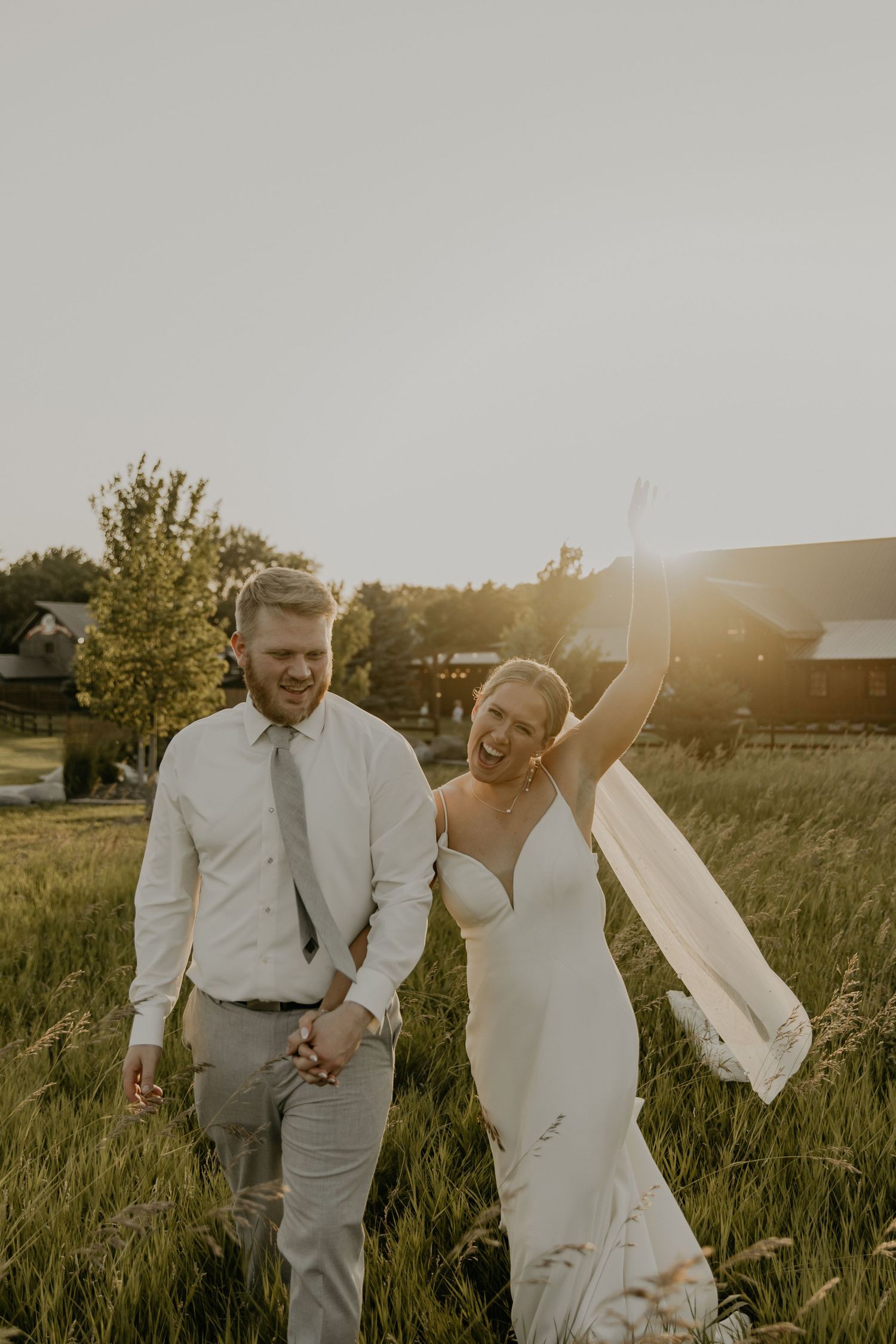 Wedding couple walks through a golden sunlit field at sunset, holding hands and laughing together.
