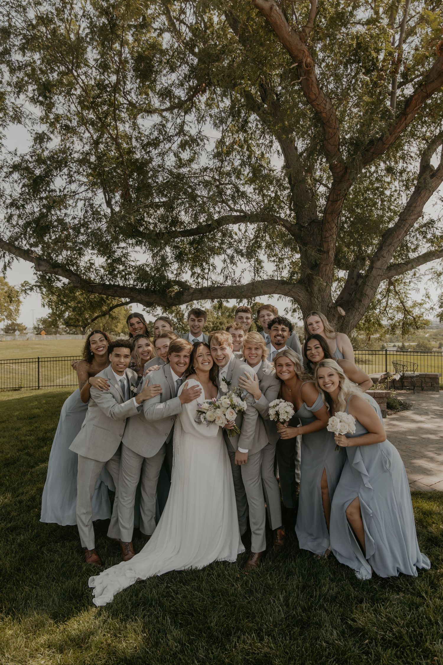 Wedding party in light gray attire poses playfully under a large tree during golden hour.