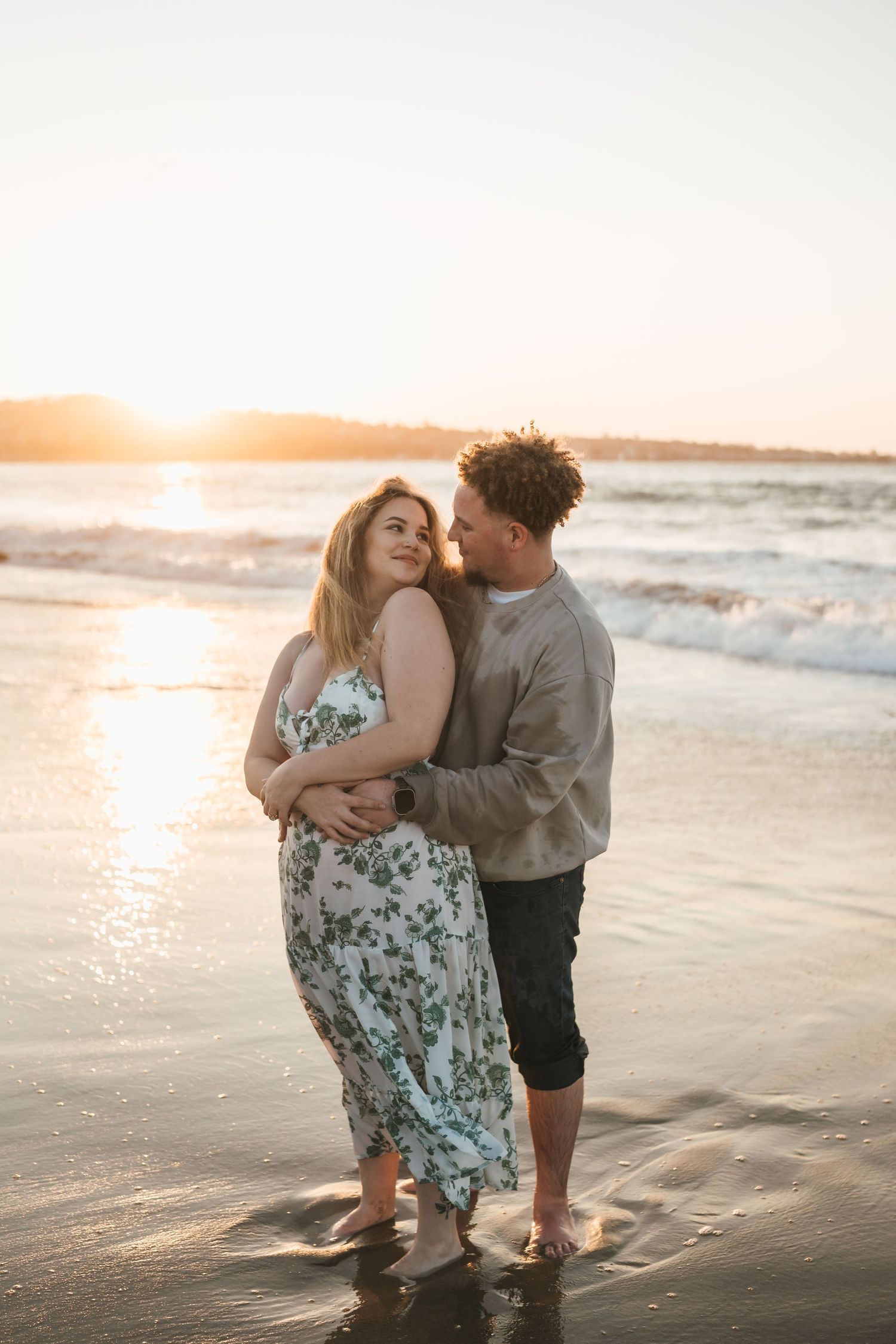 A couple embraces on a beach at sunset while waves lap at their feet and golden light illuminates the scene.