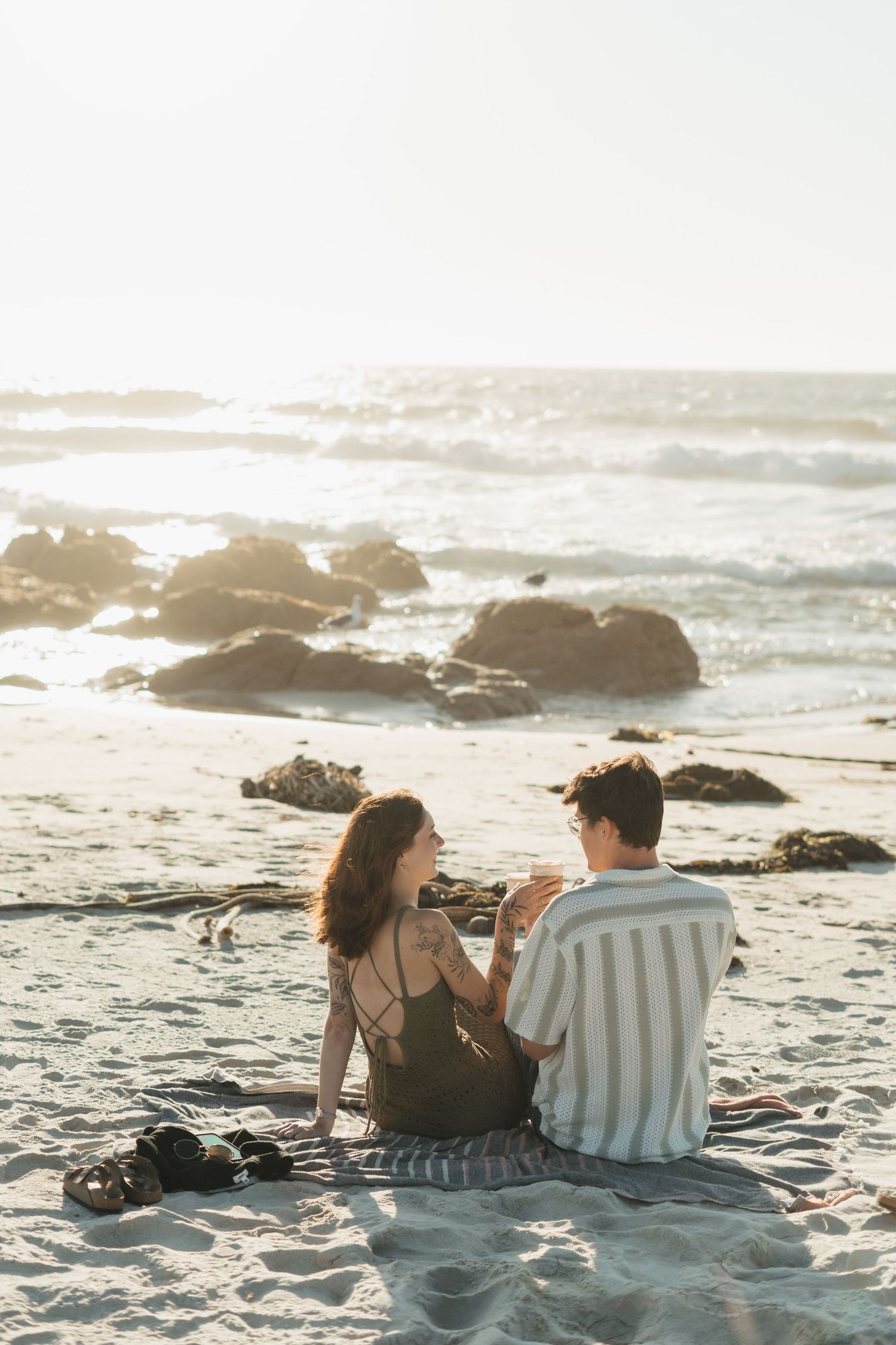 A couple sits together on a sunny beach with waves crashing against rocky outcrops during golden hour.
