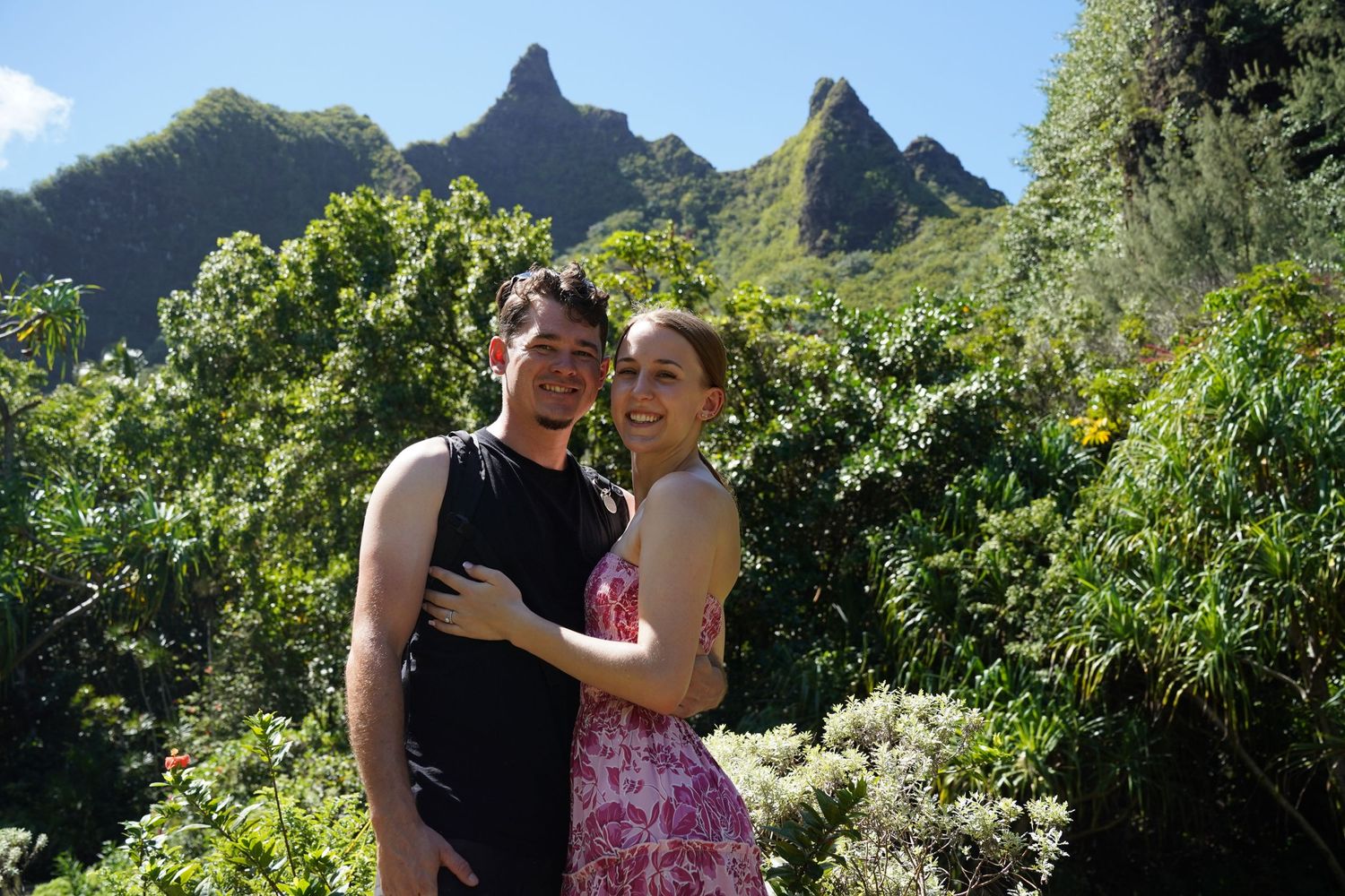 Two people pose together on a scenic hiking trail with dramatic mountain peaks in Kauai's lush landscape.