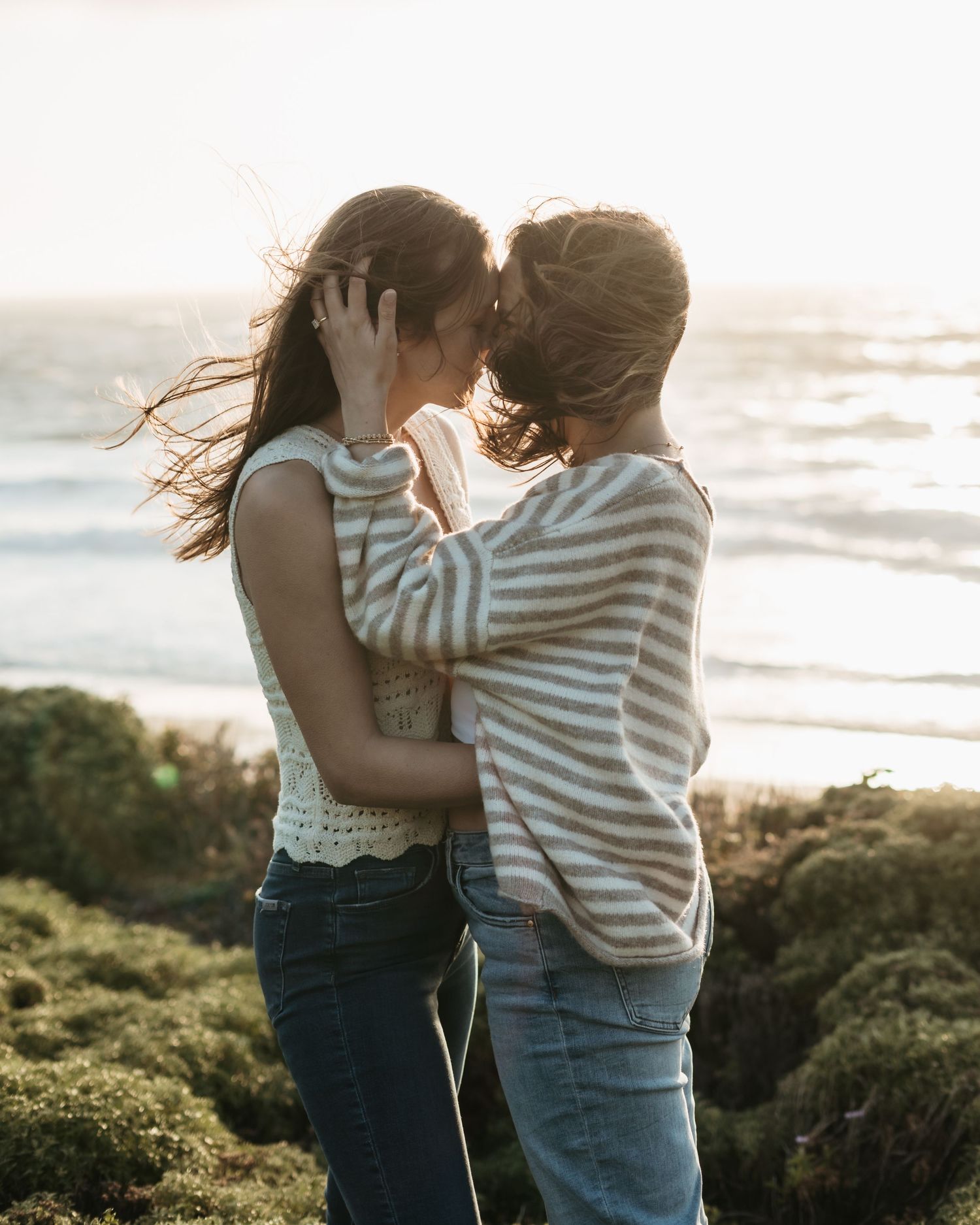 Couple sharing a romantic moment near the ocean at sunset, embracing on a coastal cliff with waves in the background.