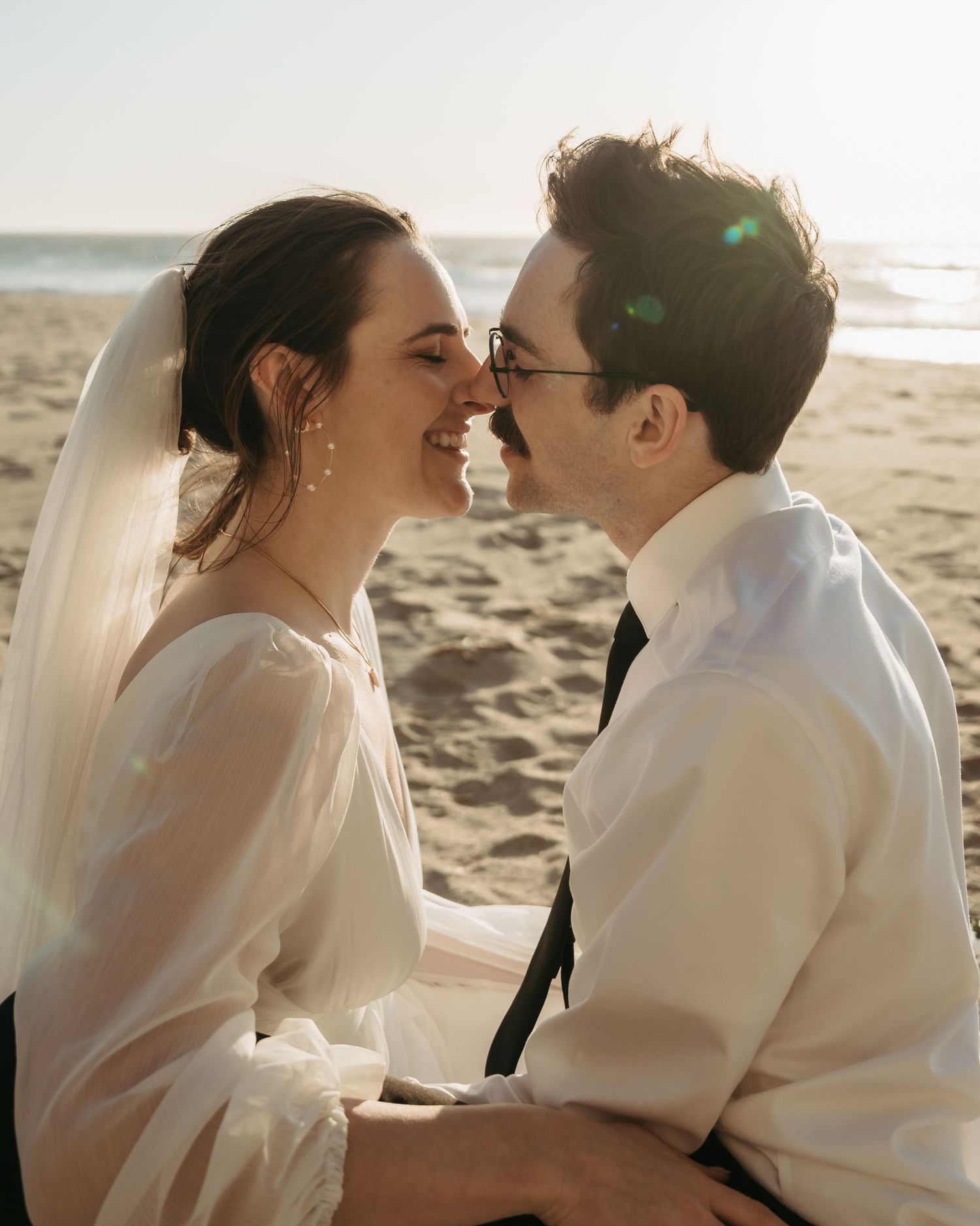 A joyful bride and groom share an intimate moment on a sunlit beach, touching noses and smiling in wedding attire.