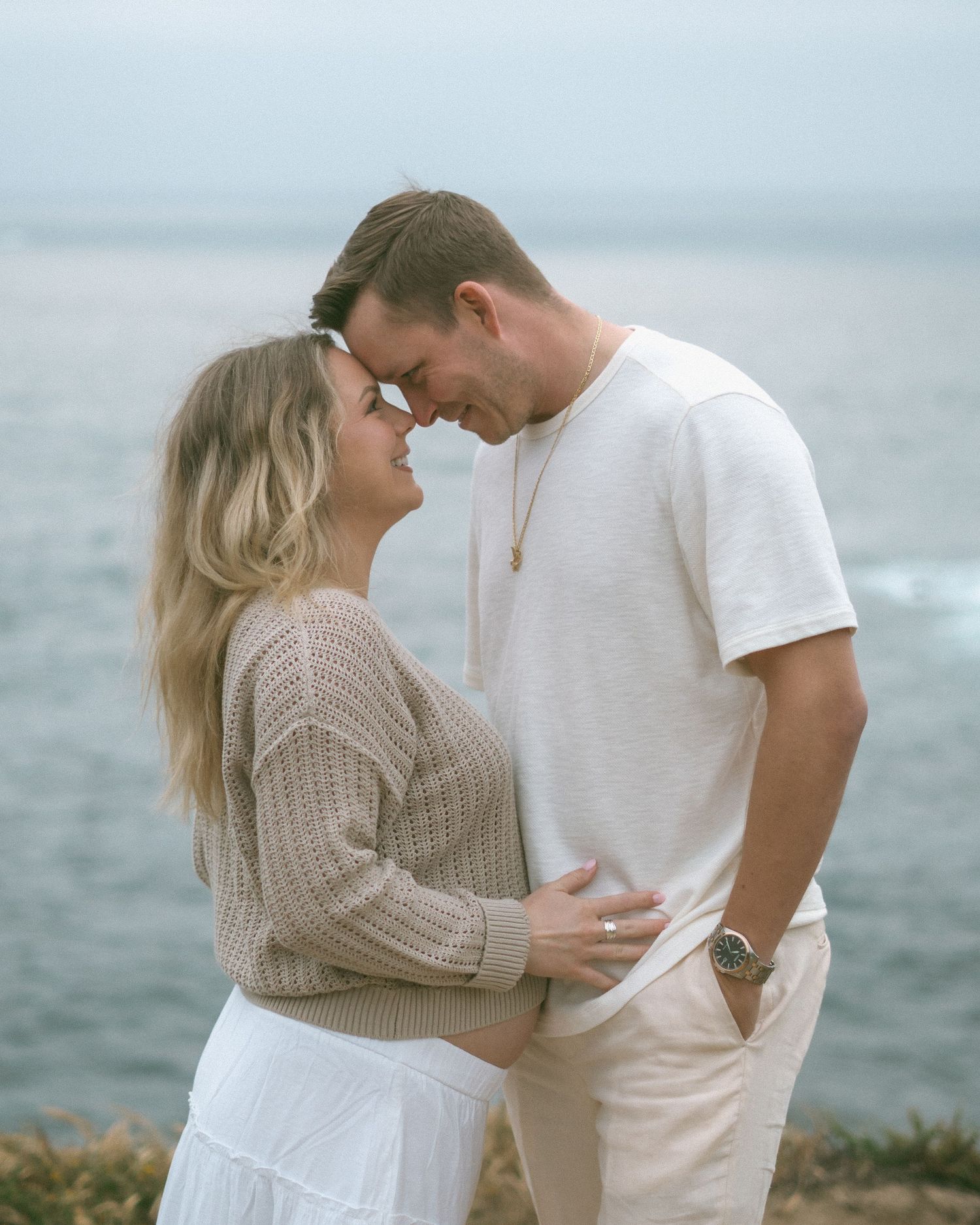 A couple shares a romantic moment by the ocean, touching foreheads and smiling, dressed in neutral tones.
