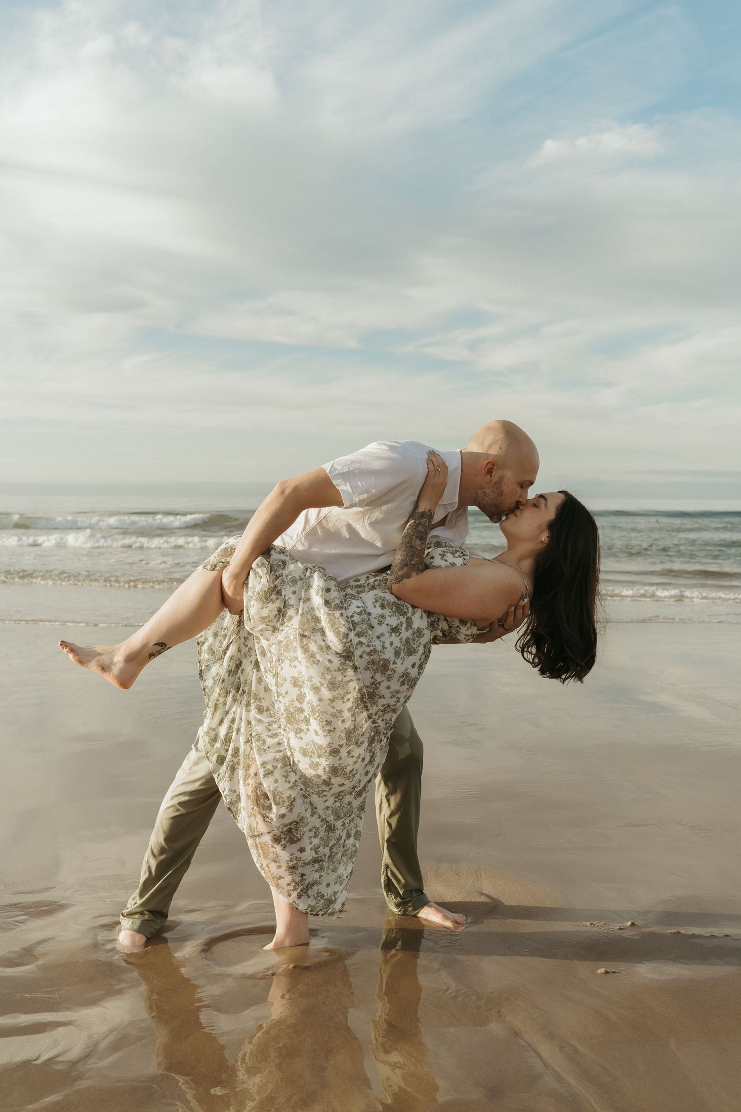 A romantic couple dancing and embracing on a sandy beach with ocean waves and cloudy sky in the background.