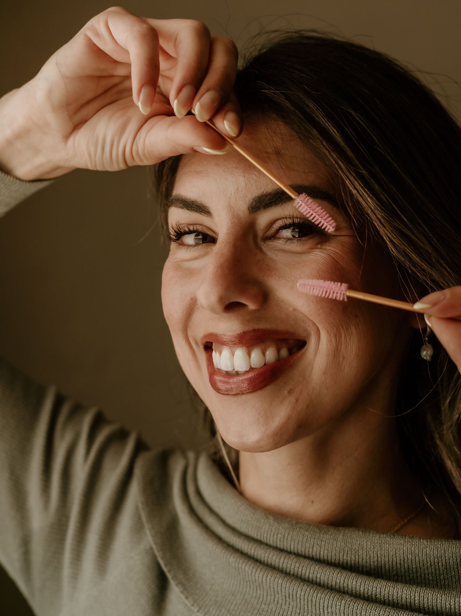 Person wearing a neutral sweater smiles warmly while grooming their eyebrows with a small brush in warm lighting.
