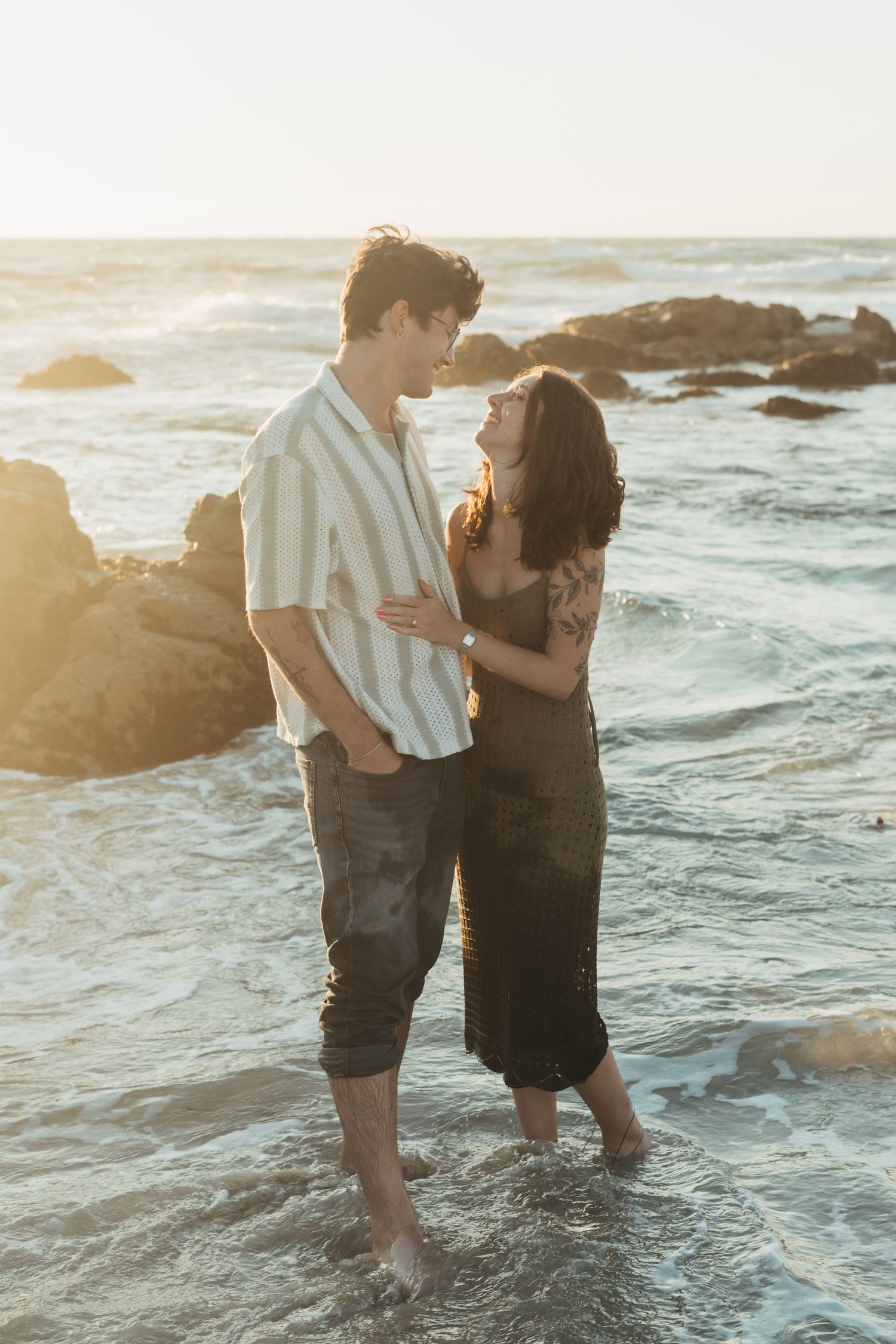 A couple embraces while standing in shallow ocean waves at sunset, creating a romantic silhouette against the water.