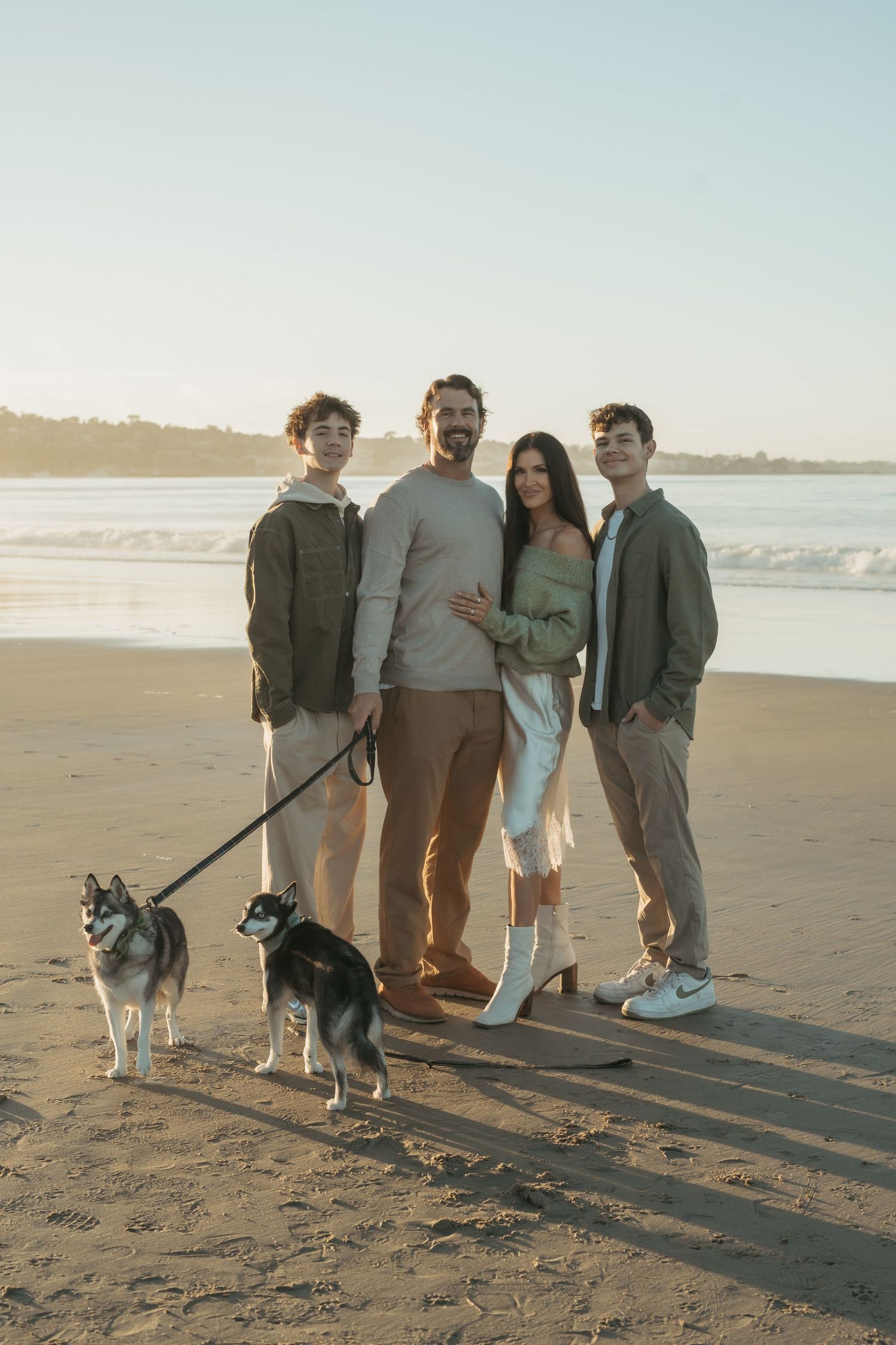 Four people and a husky dog stand together on a sandy beach at sunset casting long shadows.