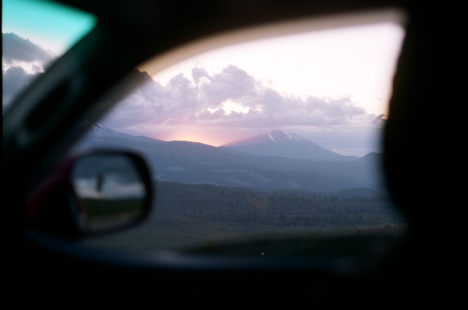 View from car window of mountain landscape at sunset with dramatic clouds and side mirror reflection.
