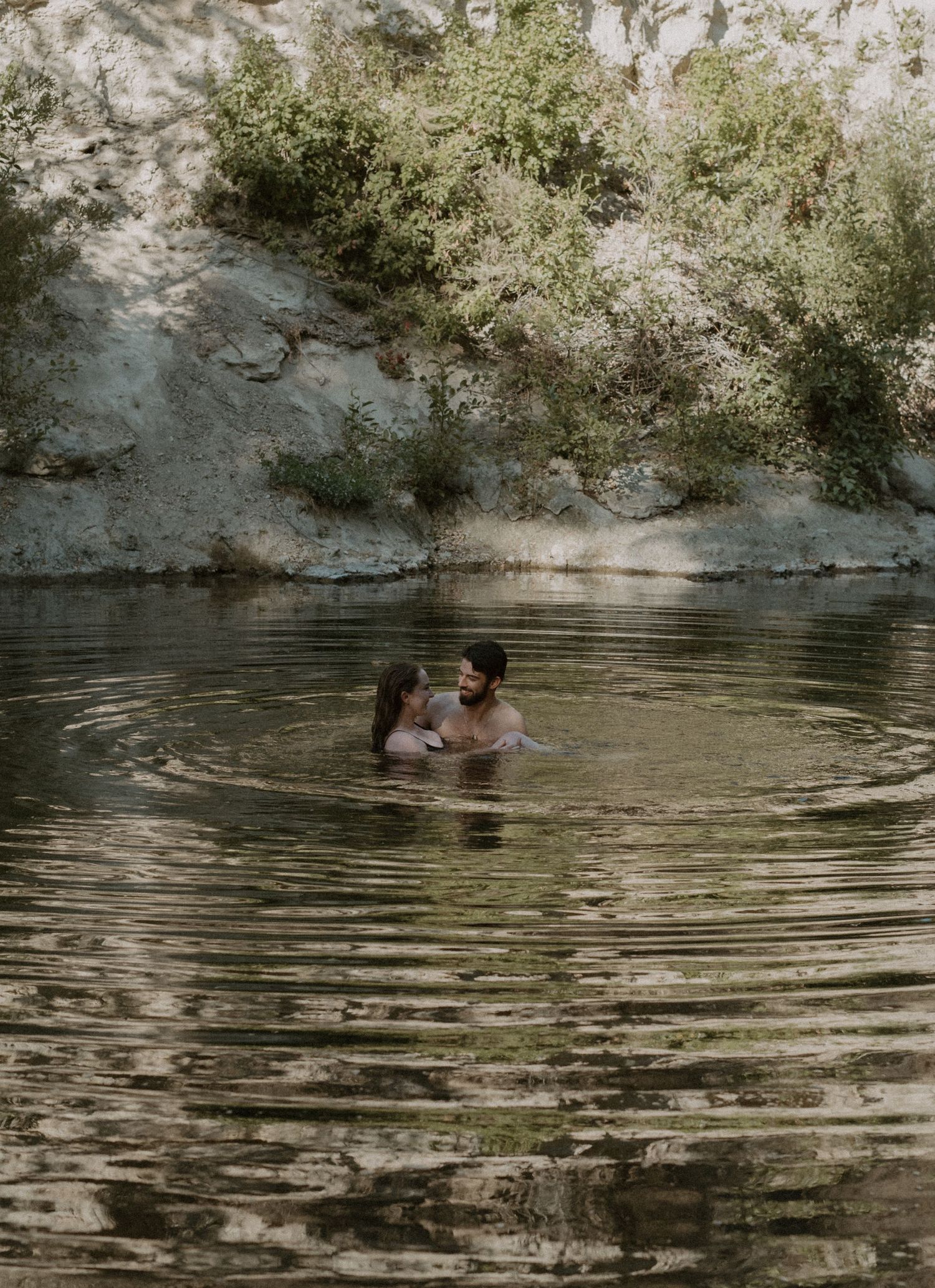 A couple embraces while wading in a tranquil swimming hole surrounded by rocky cliffs and lush greenery.
