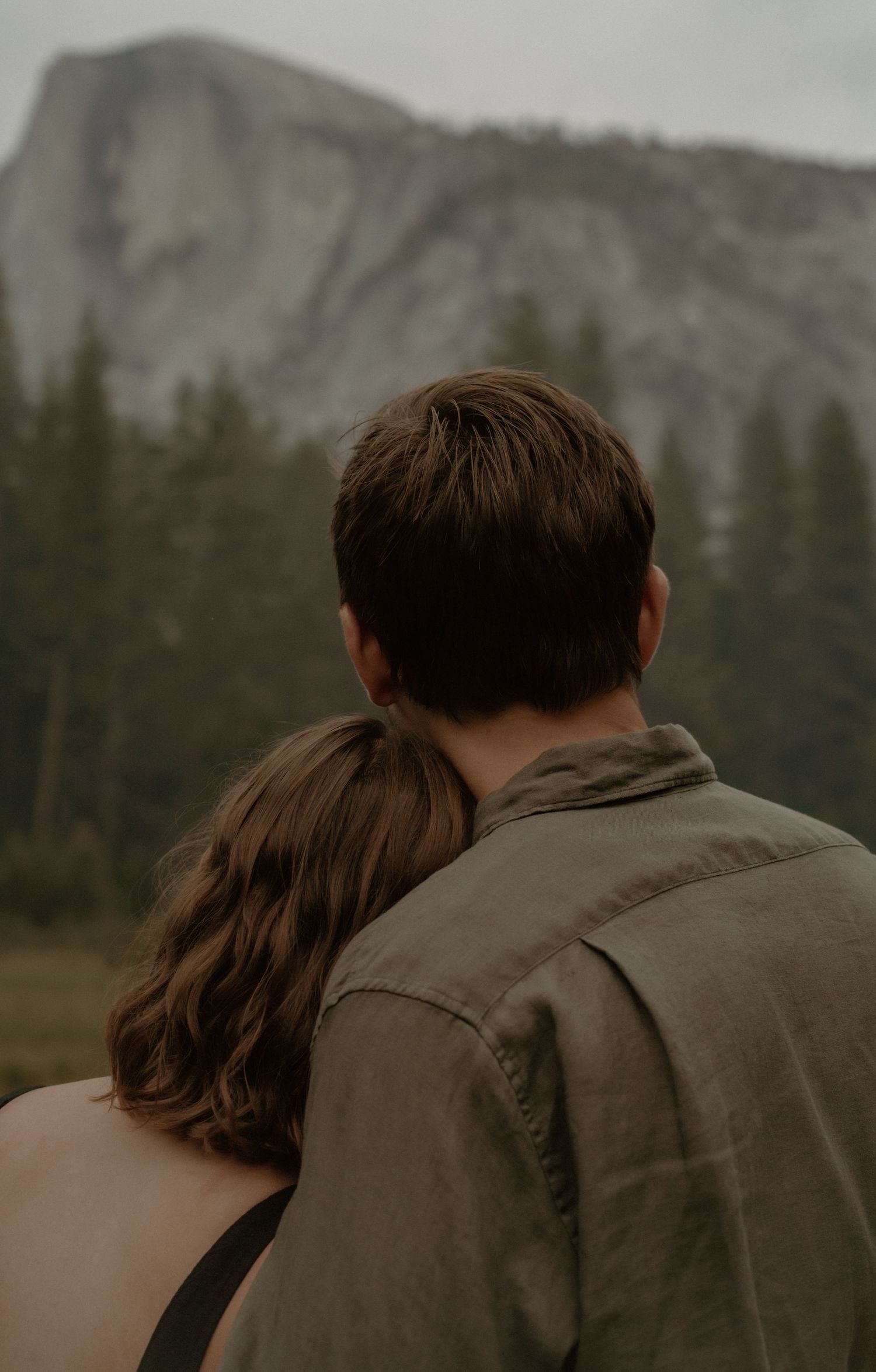 A couple in olive jackets stands close together against a mountainous backdrop in nature.