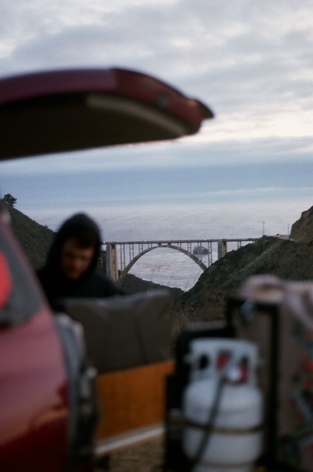 Person in hoodie near open car trunk with propane tank, coastal bridge and ocean visible in misty background.