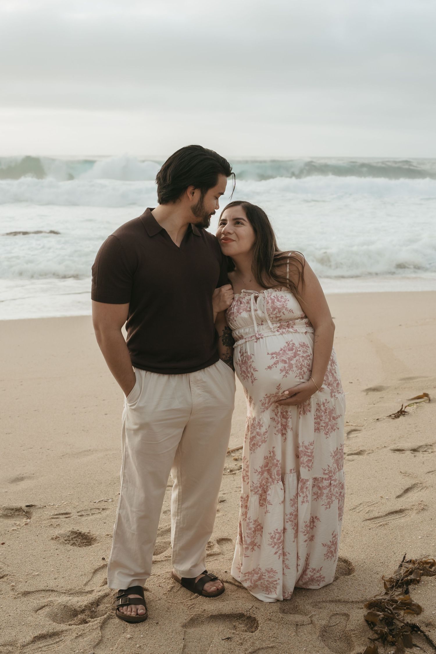 A pregnant woman and her partner embrace on a beach with crashing waves in the background.