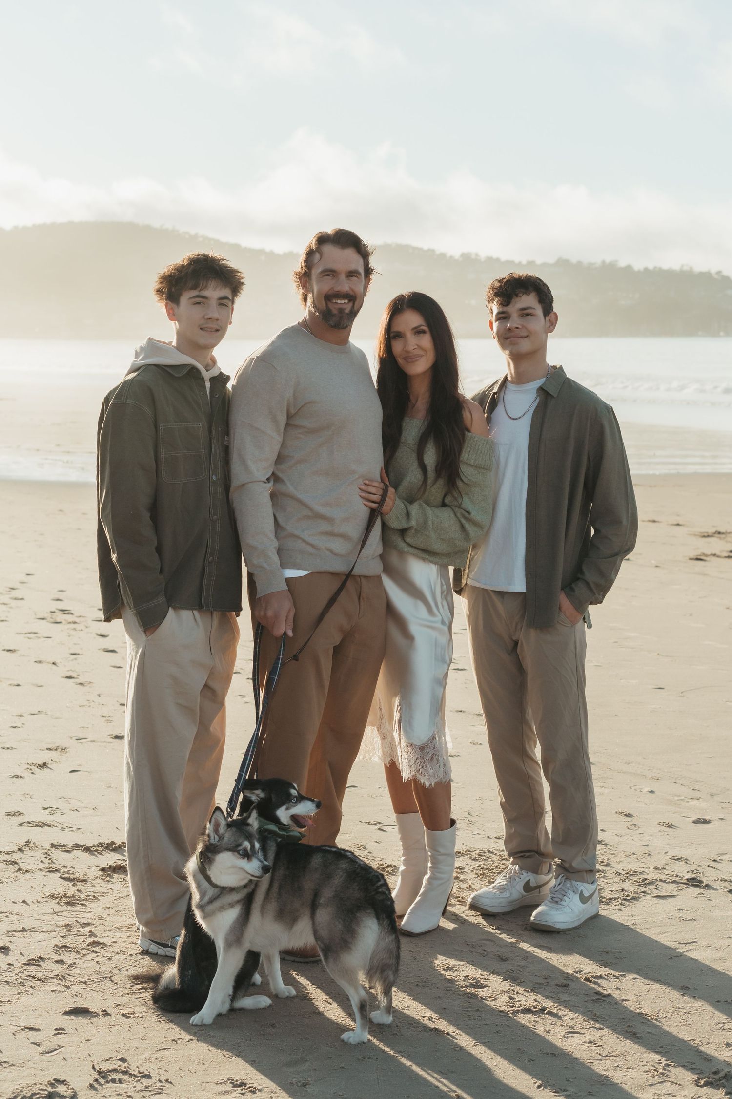 A group in casual attire stands together with their dog on a sunny beach at sunset.