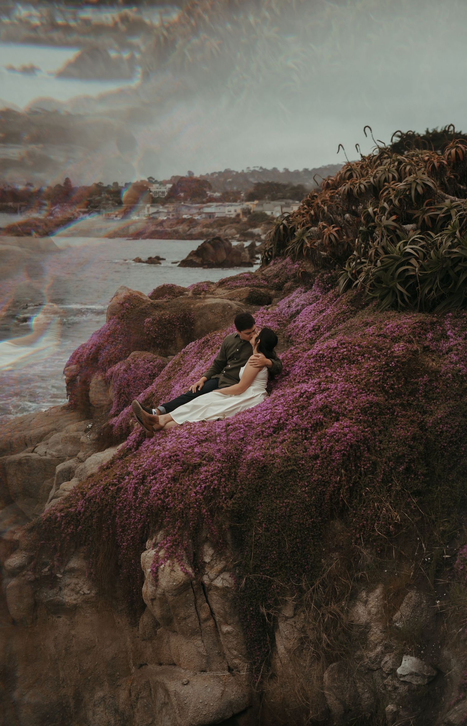 Couple sits on coastal cliff overlooking ocean with vibrant pink wildflowers blooming against moody sunset sky.