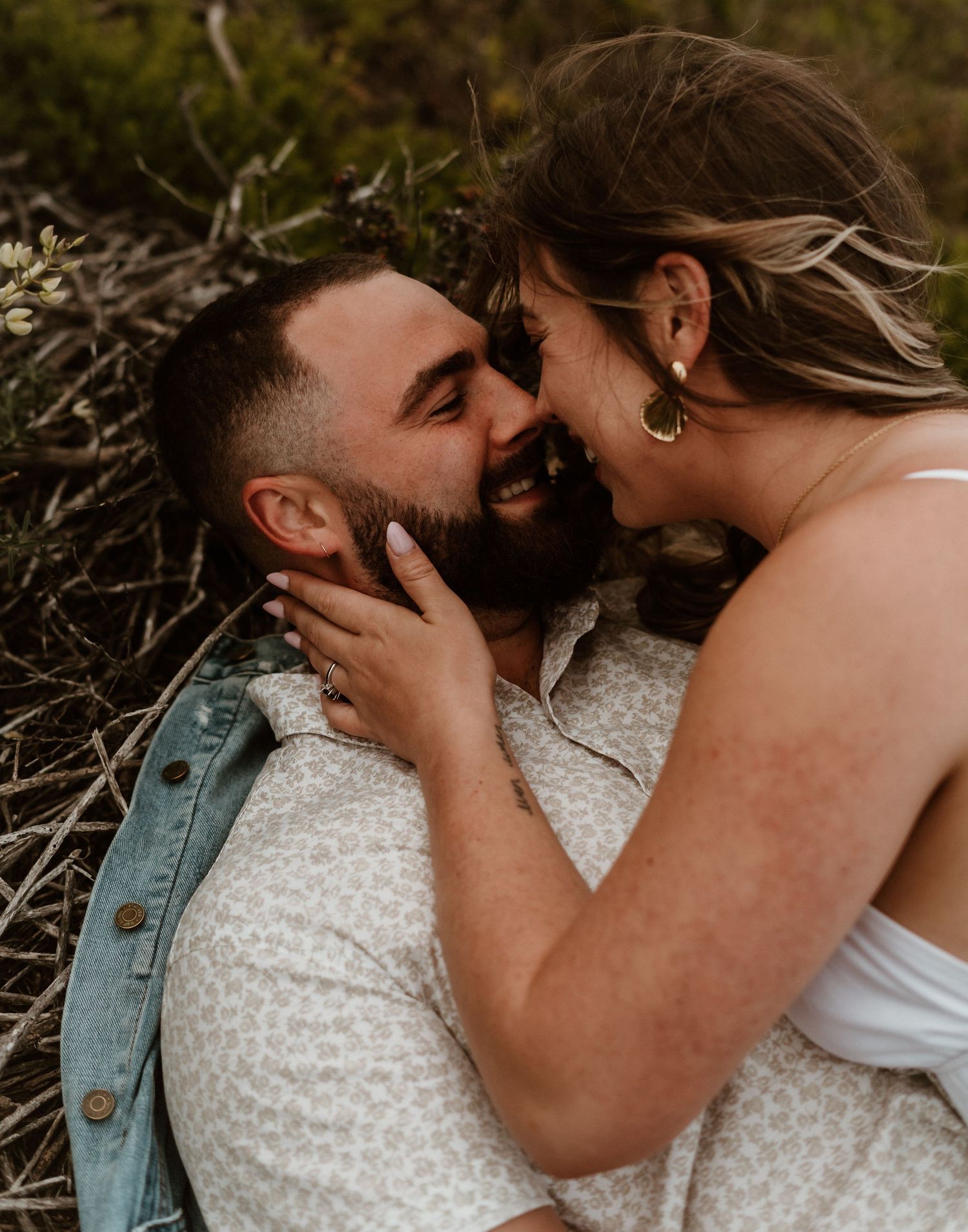 A romantic outdoor portrait showing an intimate moment between a couple laying in dry grass on a sunny day.