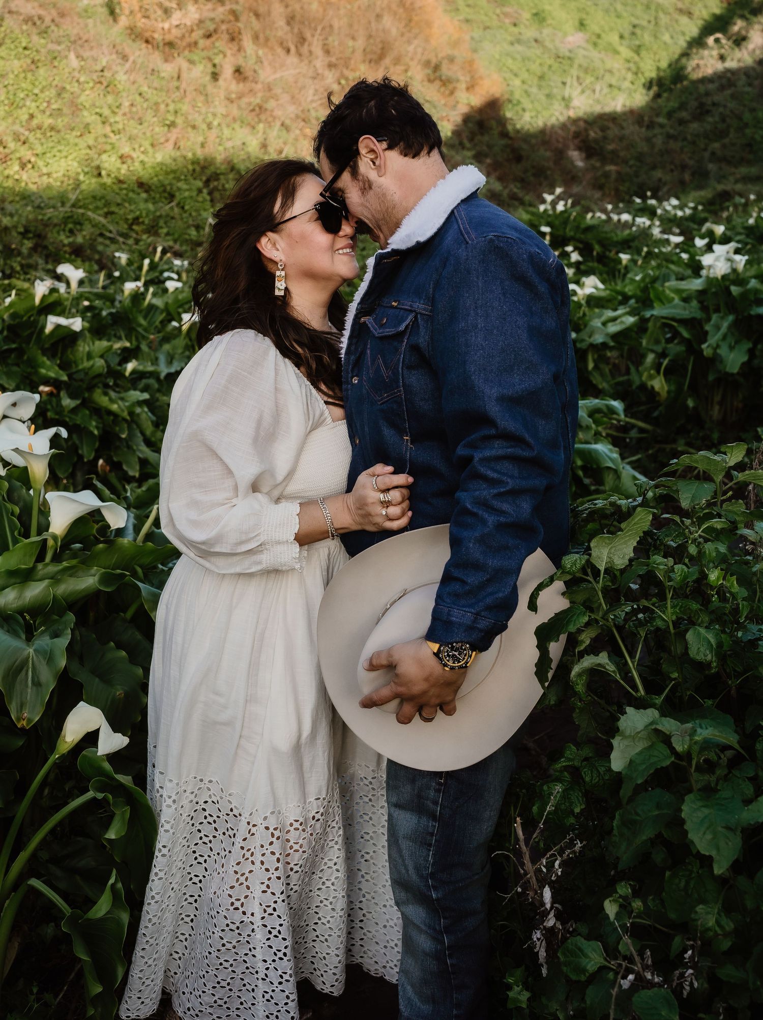Romantic couple embraces in a garden surrounded by white flowers and lush greenery on a sunny afternoon.
