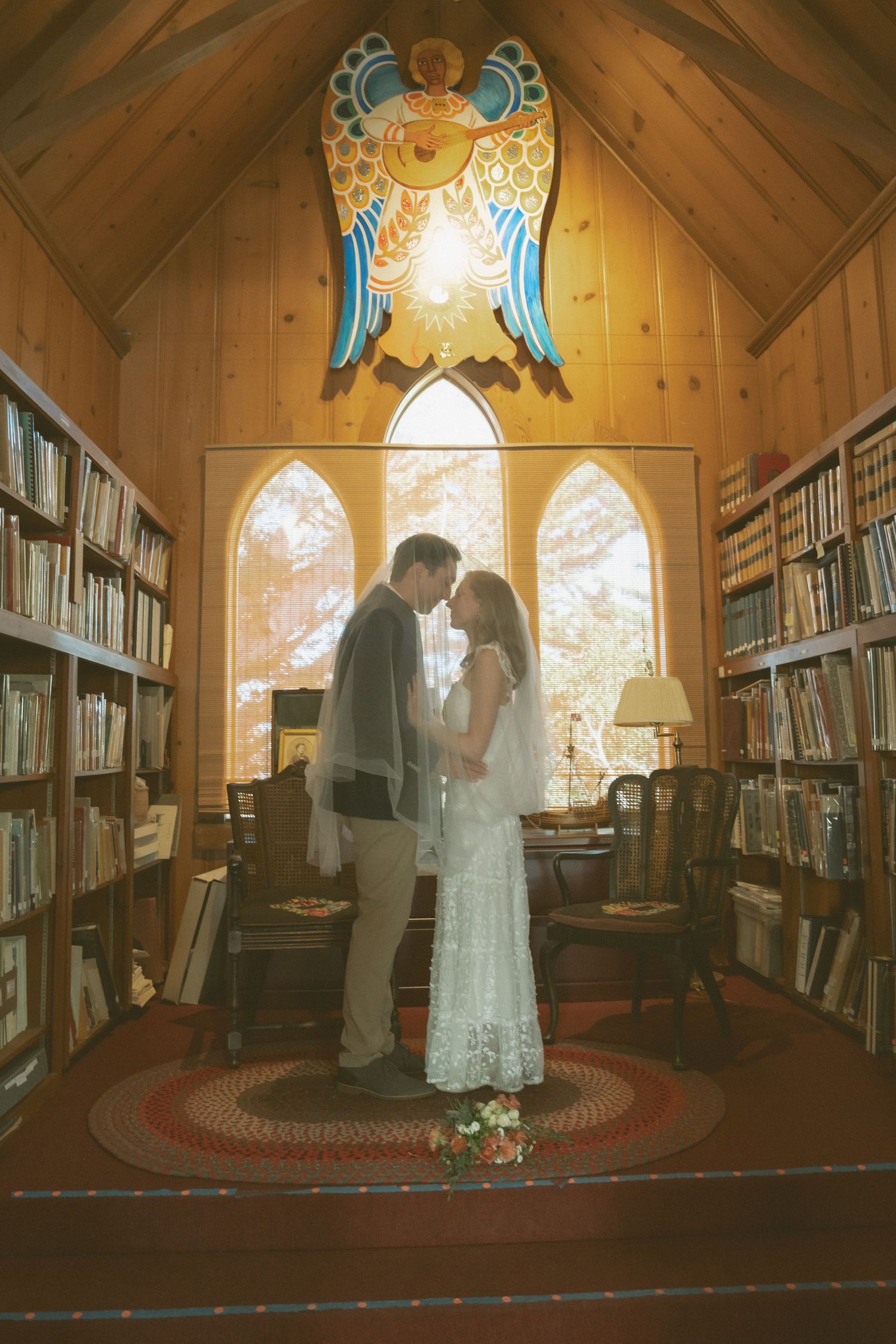 Bride and groom share romantic kiss in cozy wood-paneled library chapel with stained glass eagle and bookshelves.