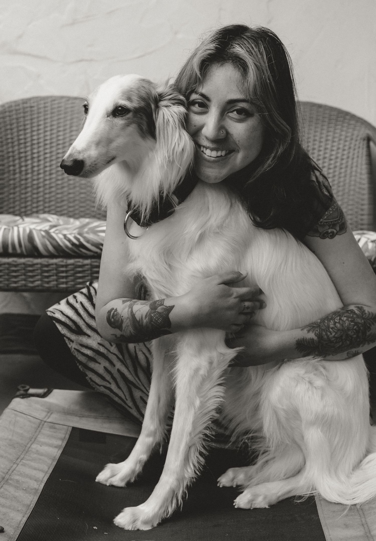 A black and white portrait of a smiling person cuddling with a beautiful Saluki or similar sighthound breed dog on a wicker chair.