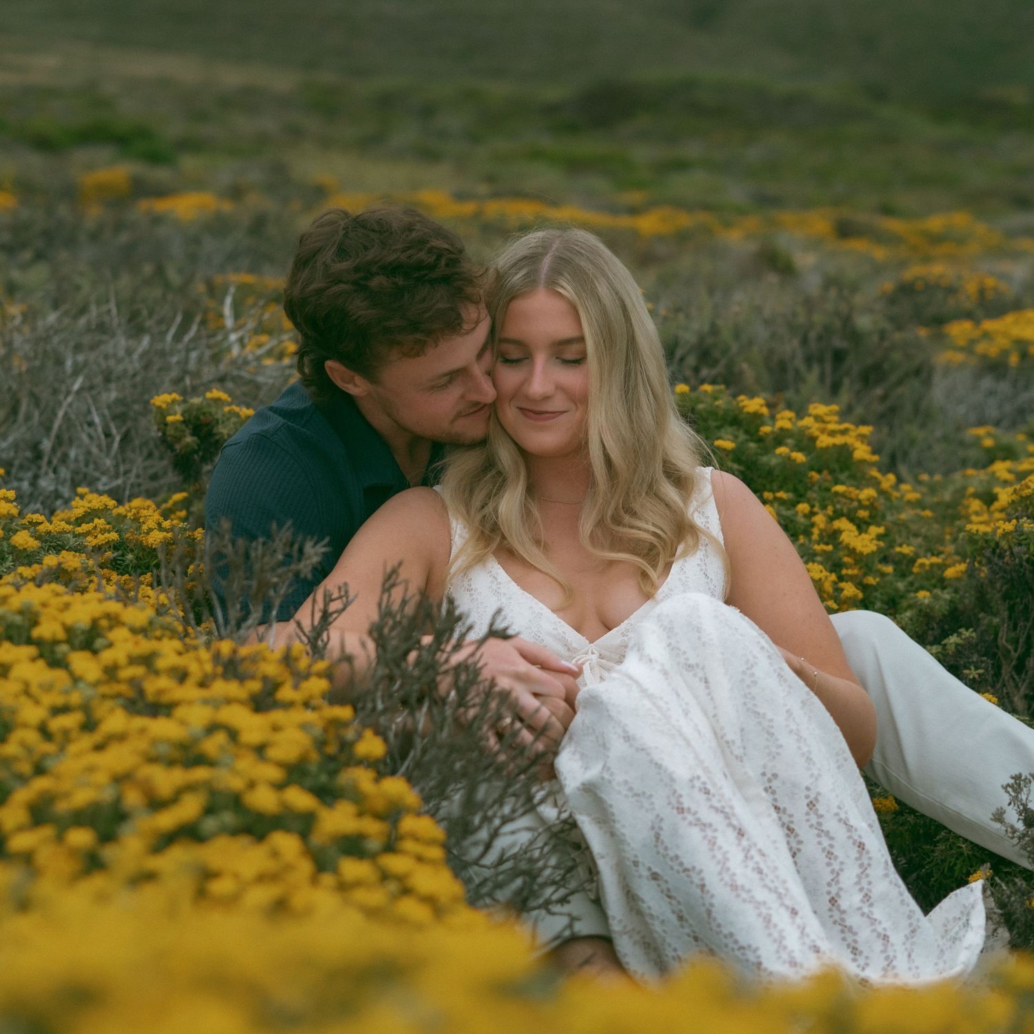 A couple sits romantically in a vibrant yellow wildflower field, the man tenderly embracing the woman in a white lace dress.