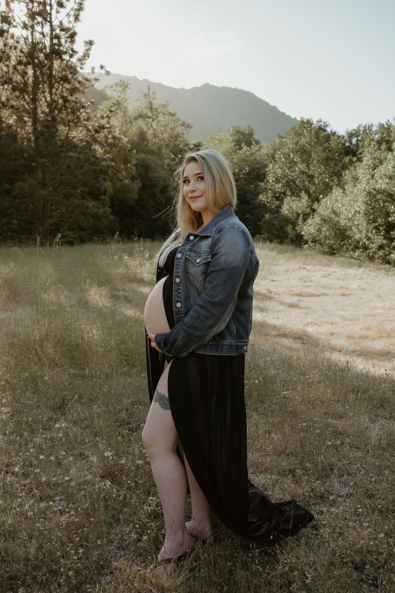 A pregnant woman in a denim jacket and black dress poses outdoors in a natural setting with mountains in the background.