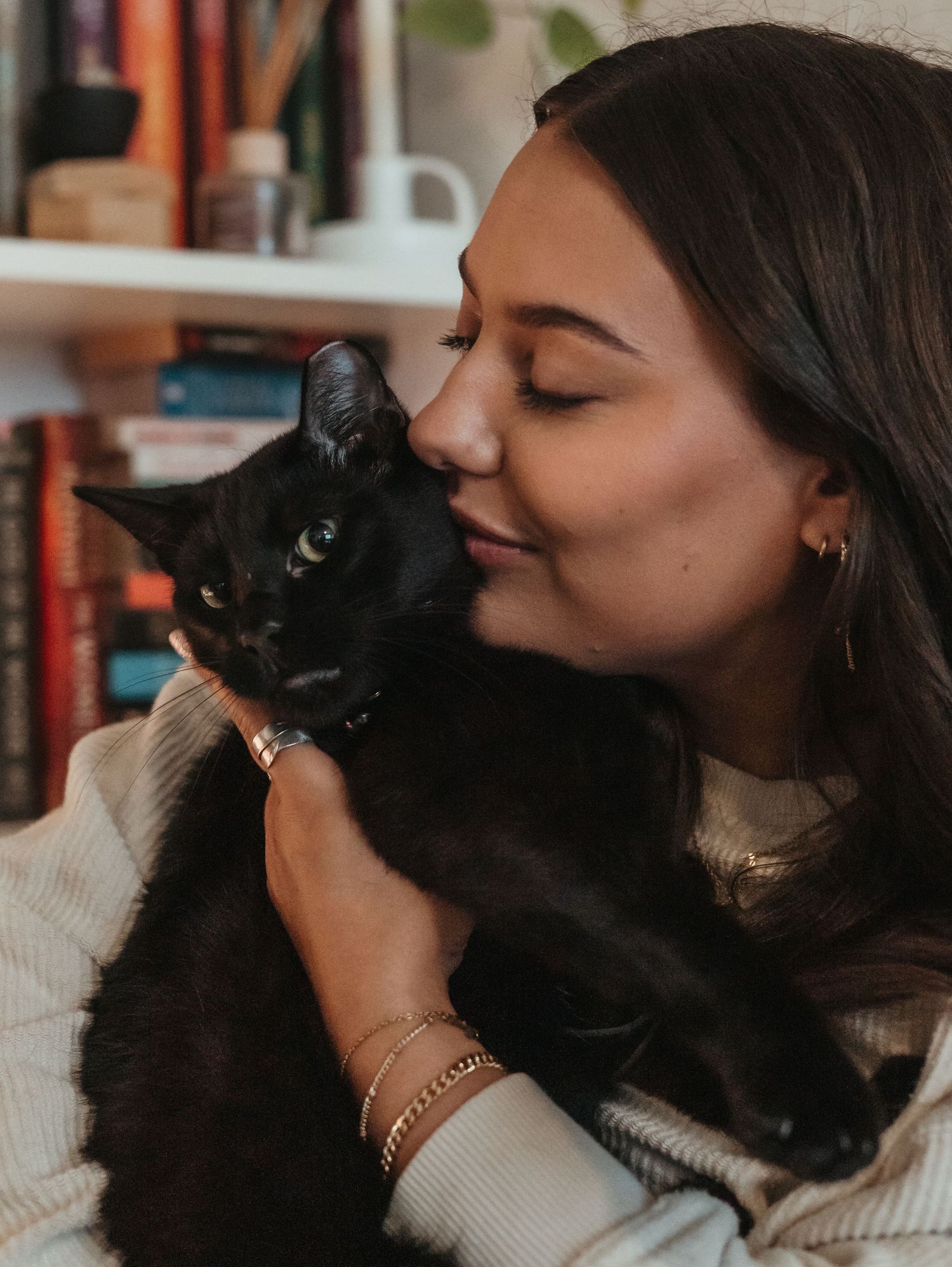 A person in a white sweater snuggles with their black cat in a cozy indoor setting.