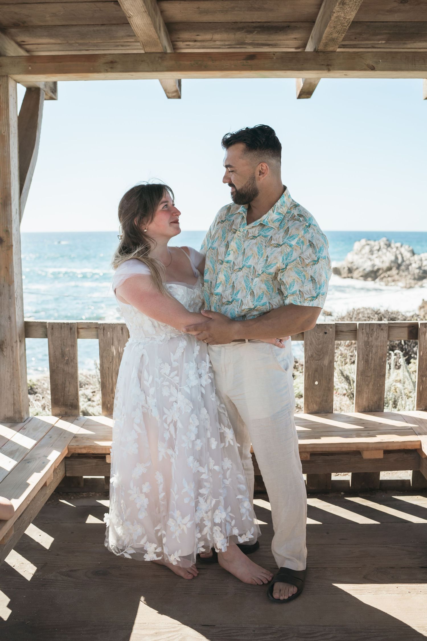 A couple shares romantic moments in a beachside gazebo wearing white and floral attire during a photo session.