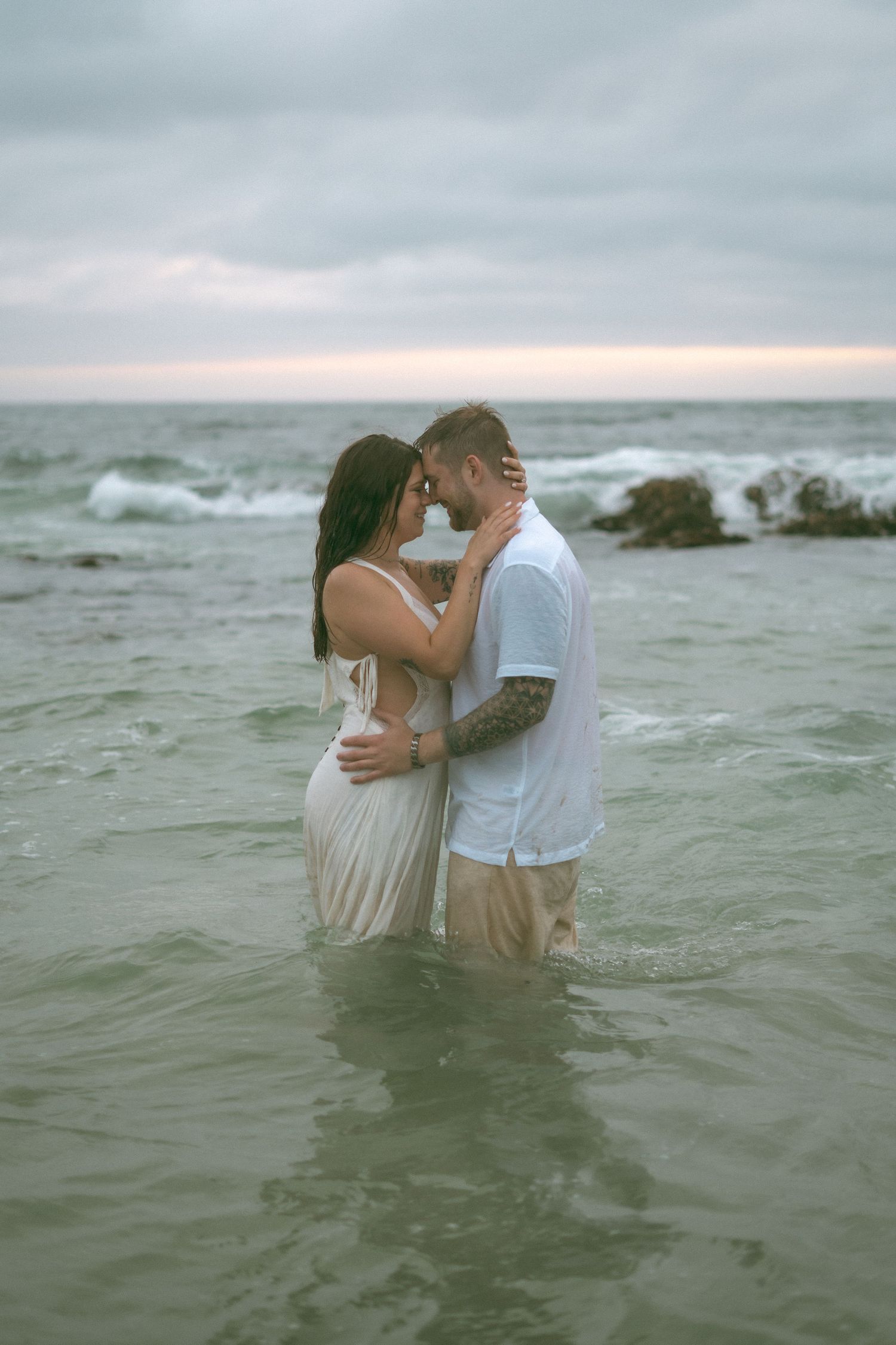 Couple embracing romantically in ocean waves at sunset, woman in white dress, man with tattoos in white shirt.