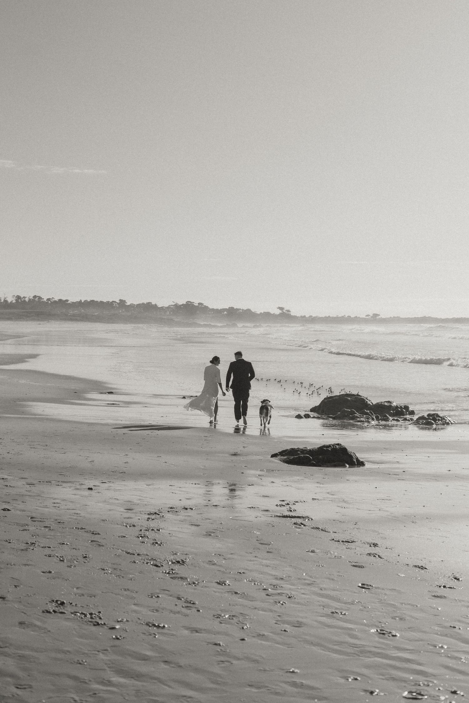 People walking along a misty beach with dark rocks scattered on the sand and ocean waves in black and white.