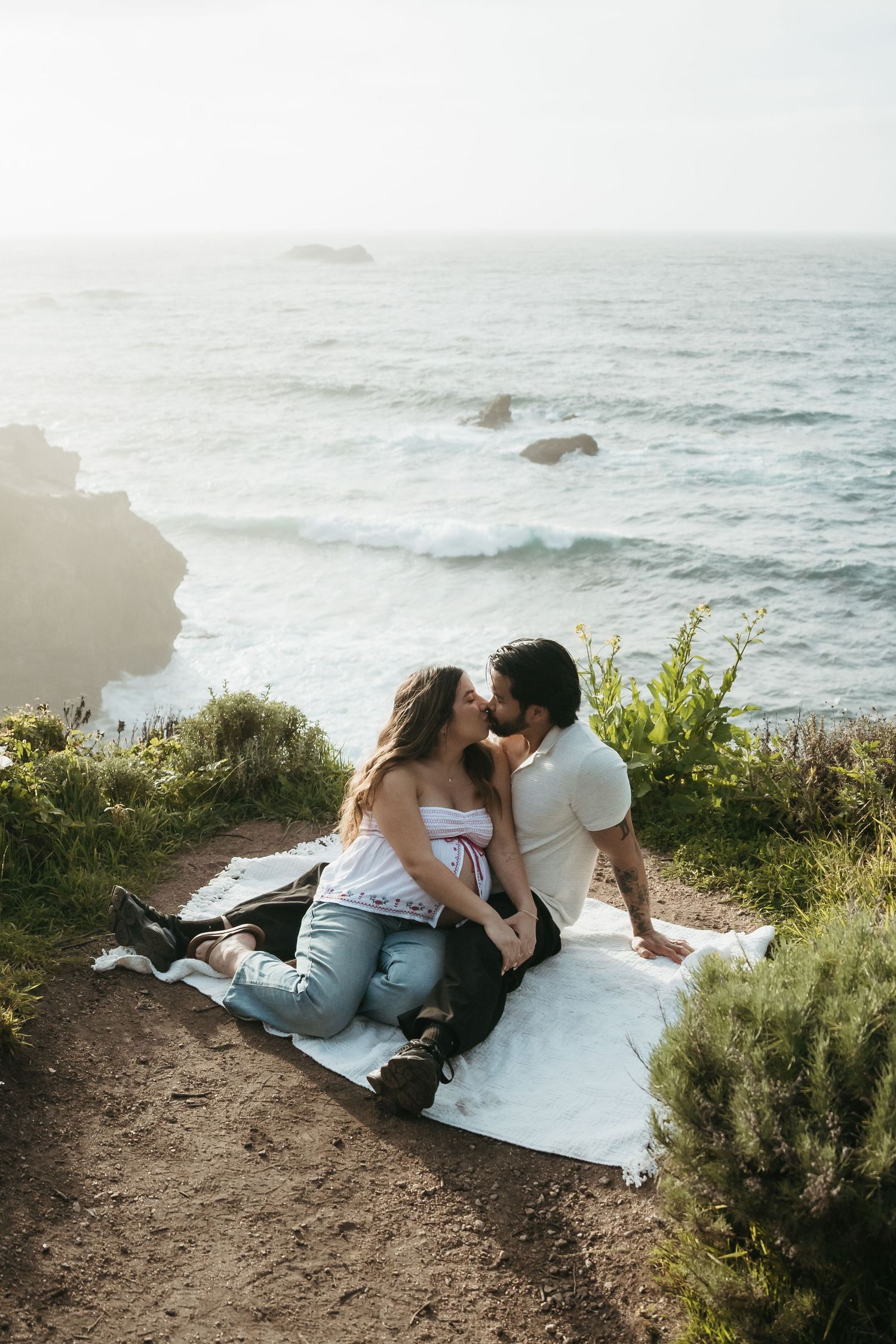 Couple sharing a romantic kiss on a blanket atop a coastal cliff with ocean waves and rocks in the background.