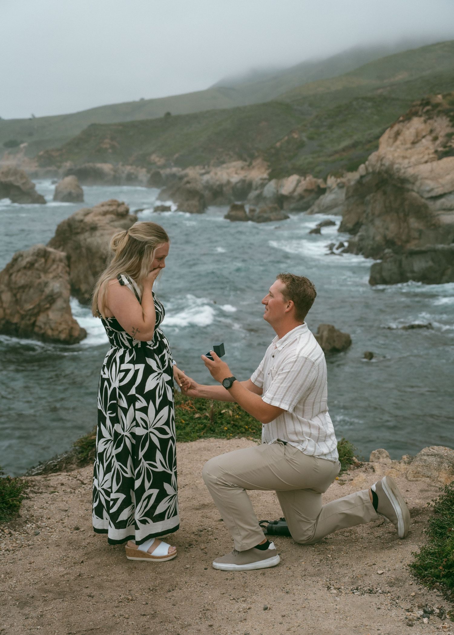 Man proposes to woman on rocky Big Sur coastline cliff, holding ring box while kneeling on scenic California ocean overlook.