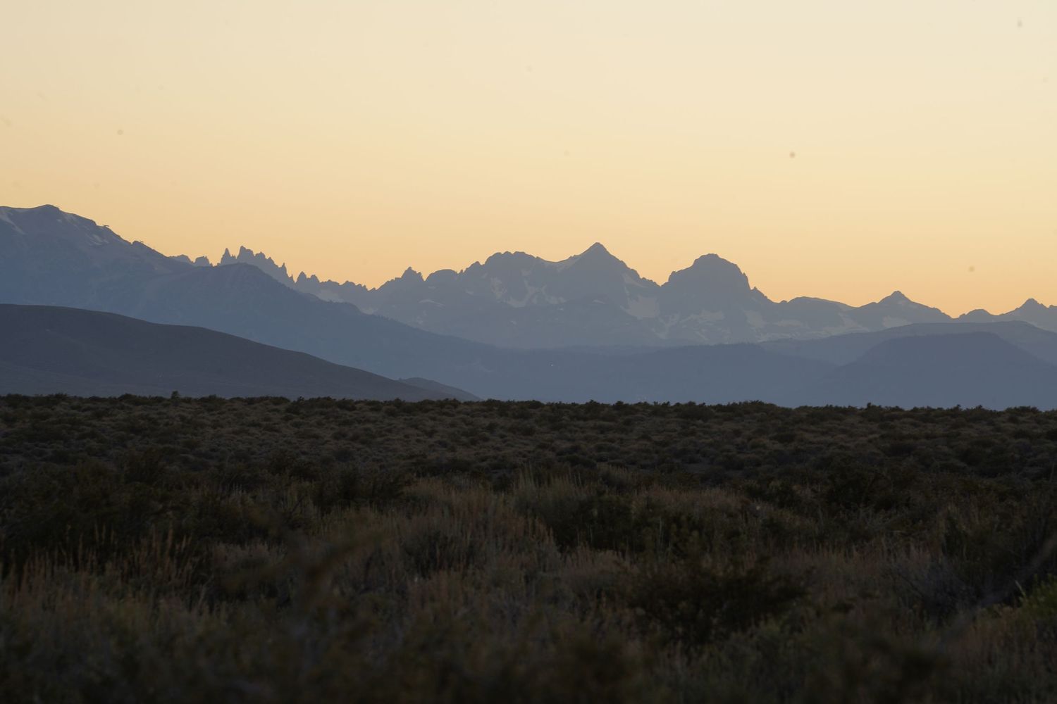 Desert scrubland at dusk with dramatic mountain silhouettes against a warm orange sunset sky.