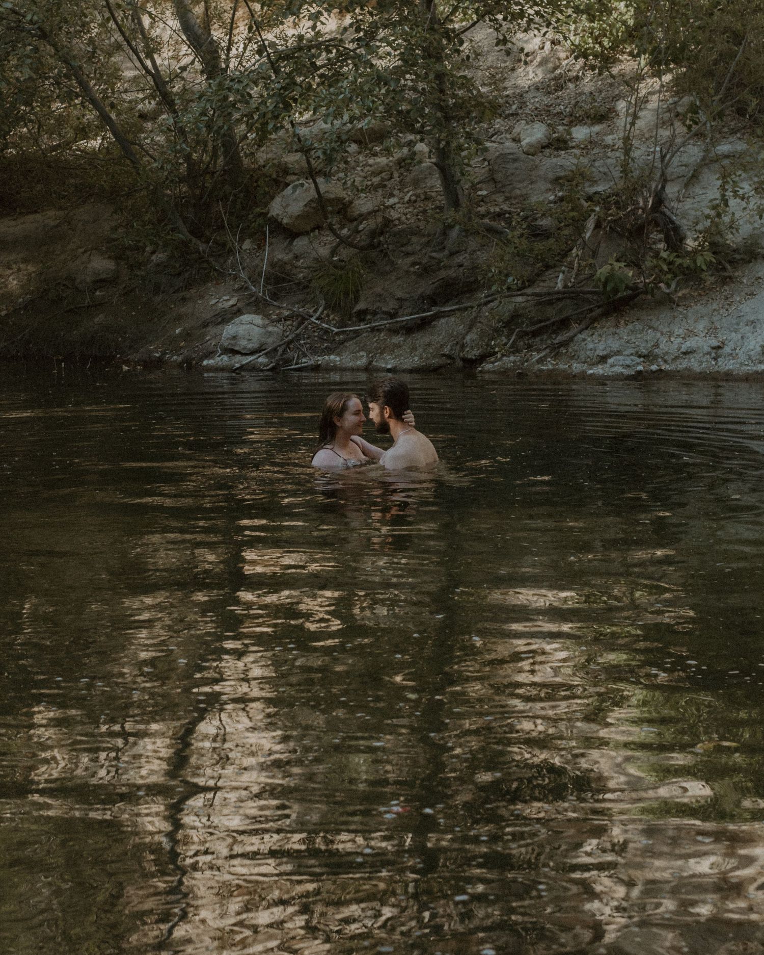 Two people share an intimate moment while swimming in a secluded forest pond with dark, reflective waters.