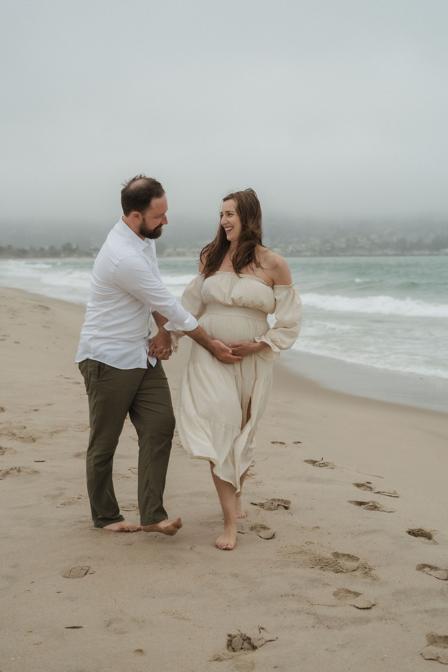 A couple shares a romantic moment while dancing together on a misty beach in soft neutral tones.