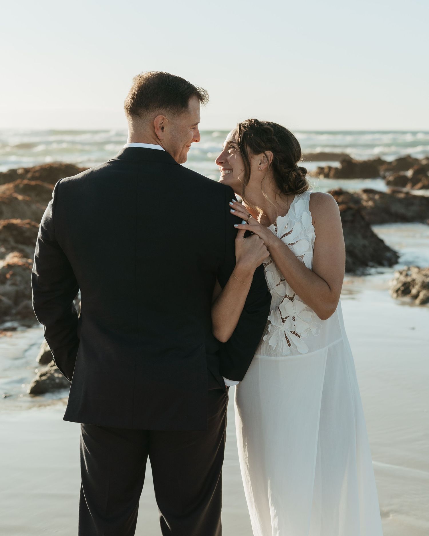 A bride and groom share an intimate moment on a rocky beach at sunset.