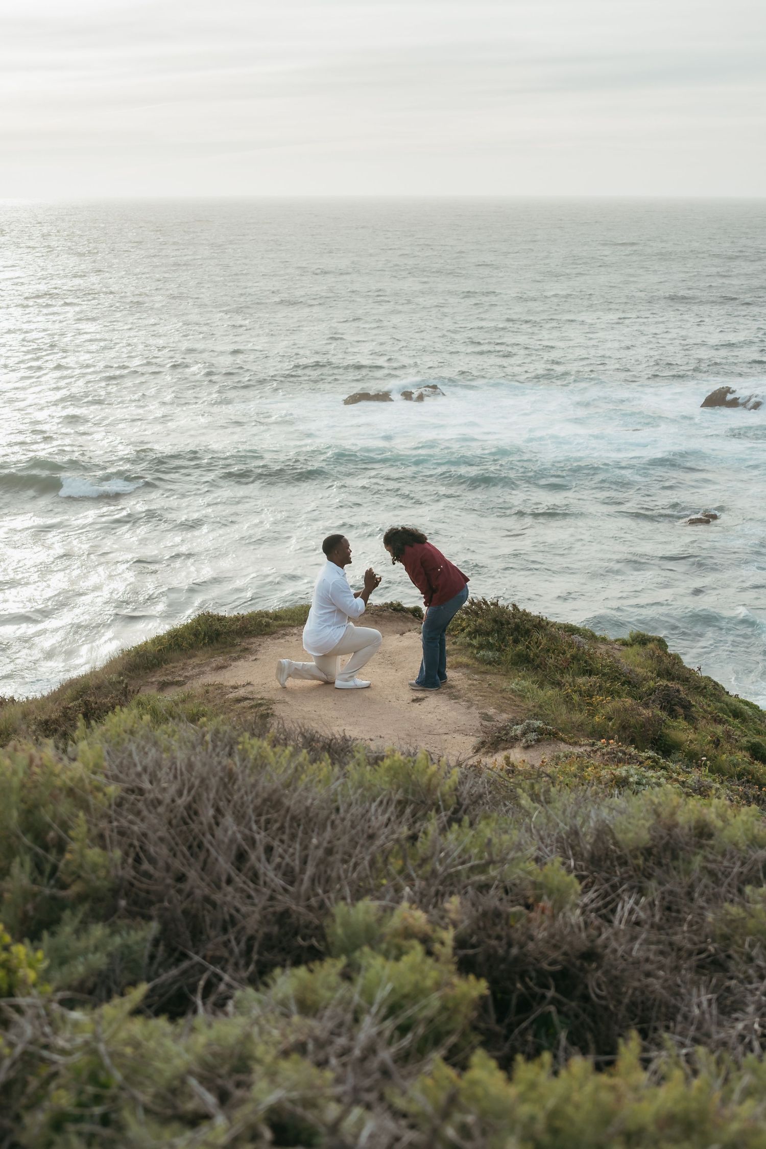 A man proposes on one knee to a woman on a scenic coastal cliff overlooking the ocean waves.