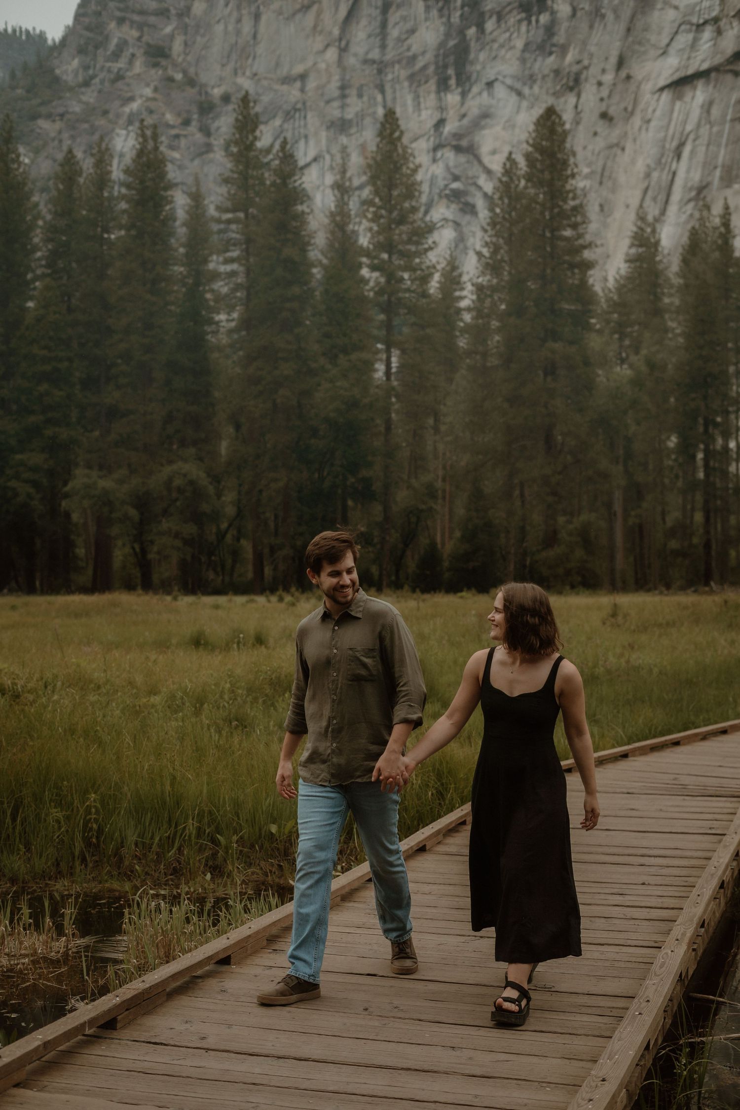 A couple walks hand in hand along a wooden boardwalk path through a scenic meadow with granite cliffs in Yosemite Valley.