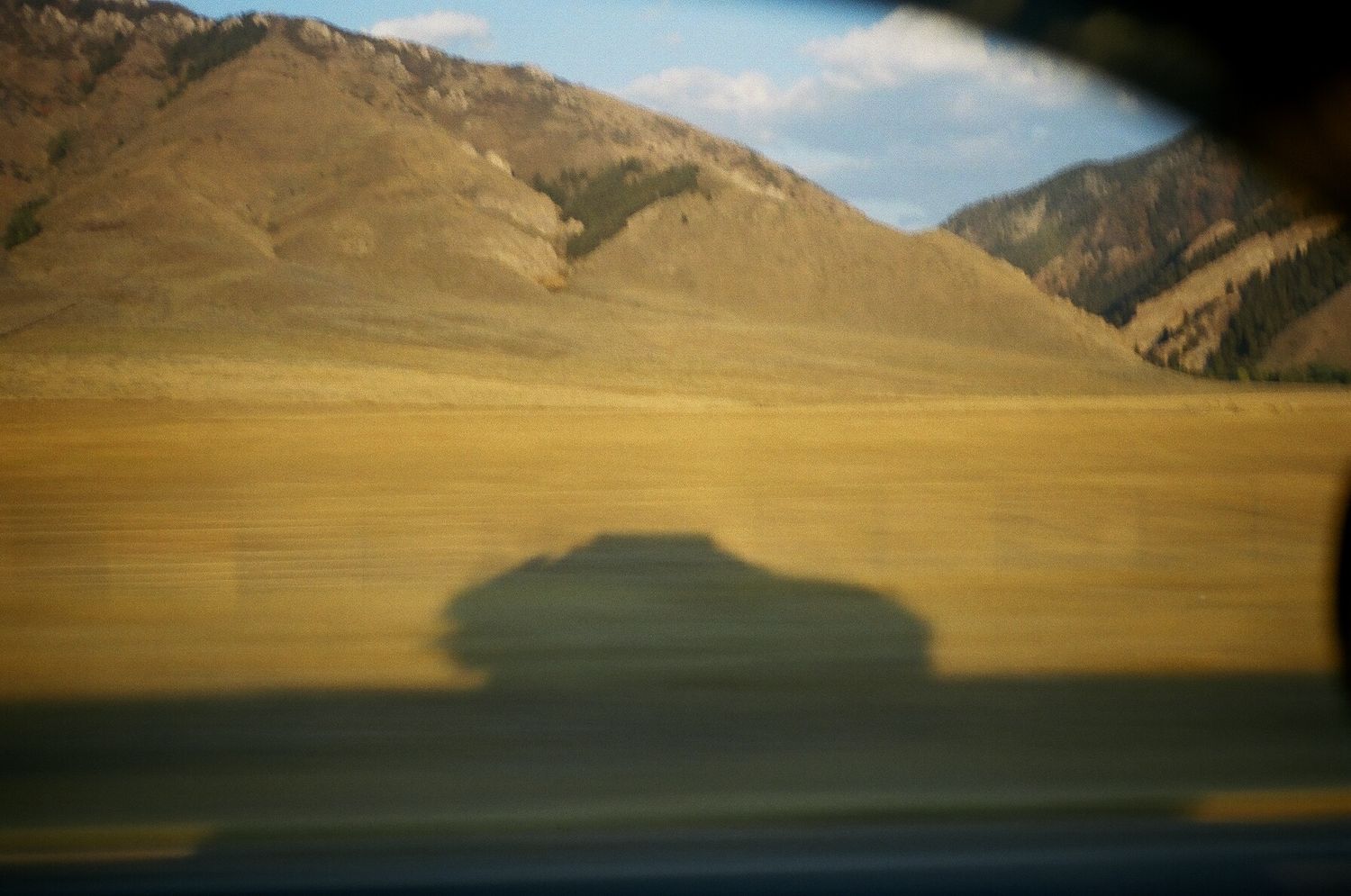 Shadow of a car cast on golden plains with mountain range backdrop, captured through a vehicle window in motion.