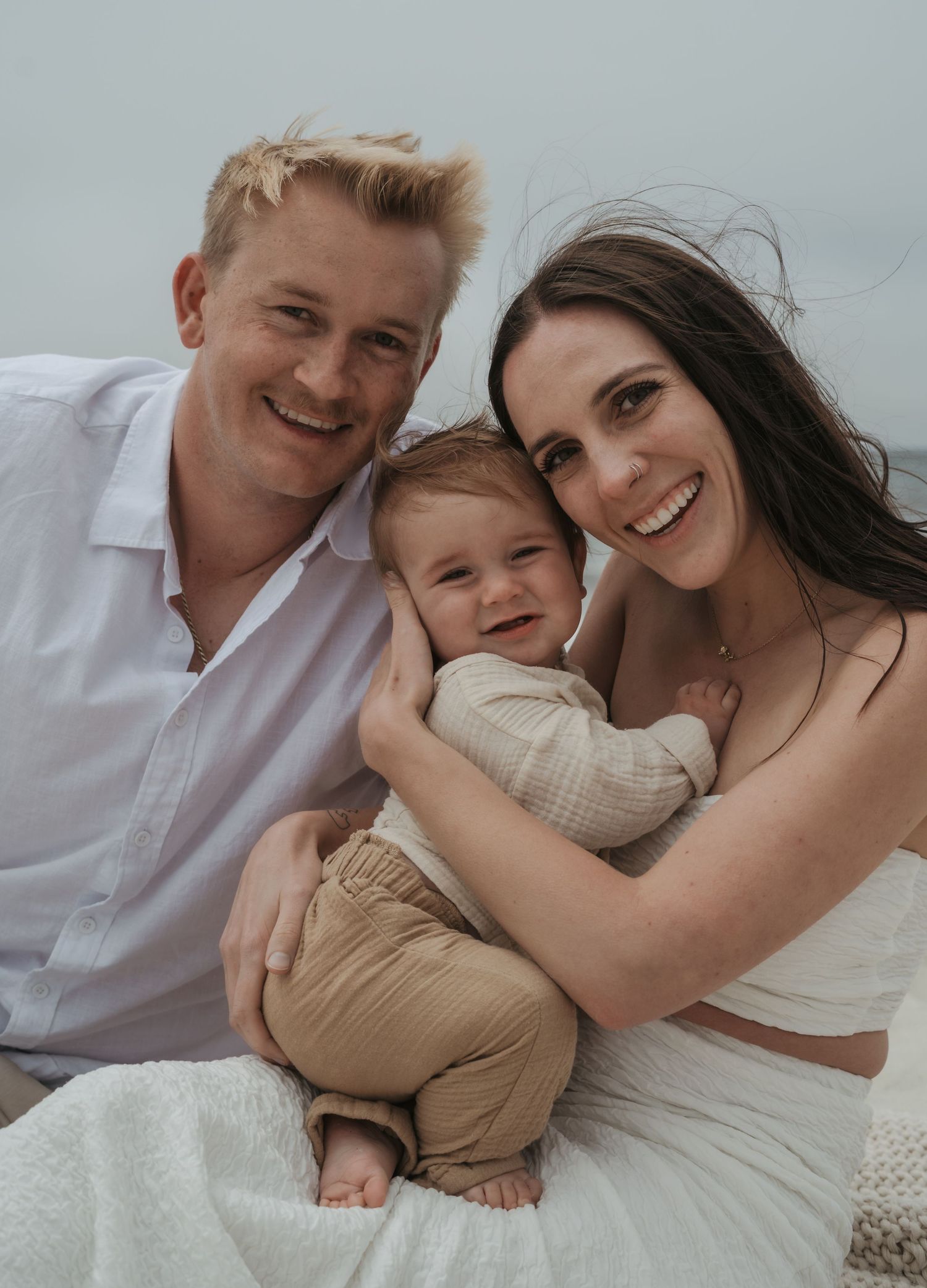 A happy family of three poses together outdoors in white and beige outfits with a neutral background.